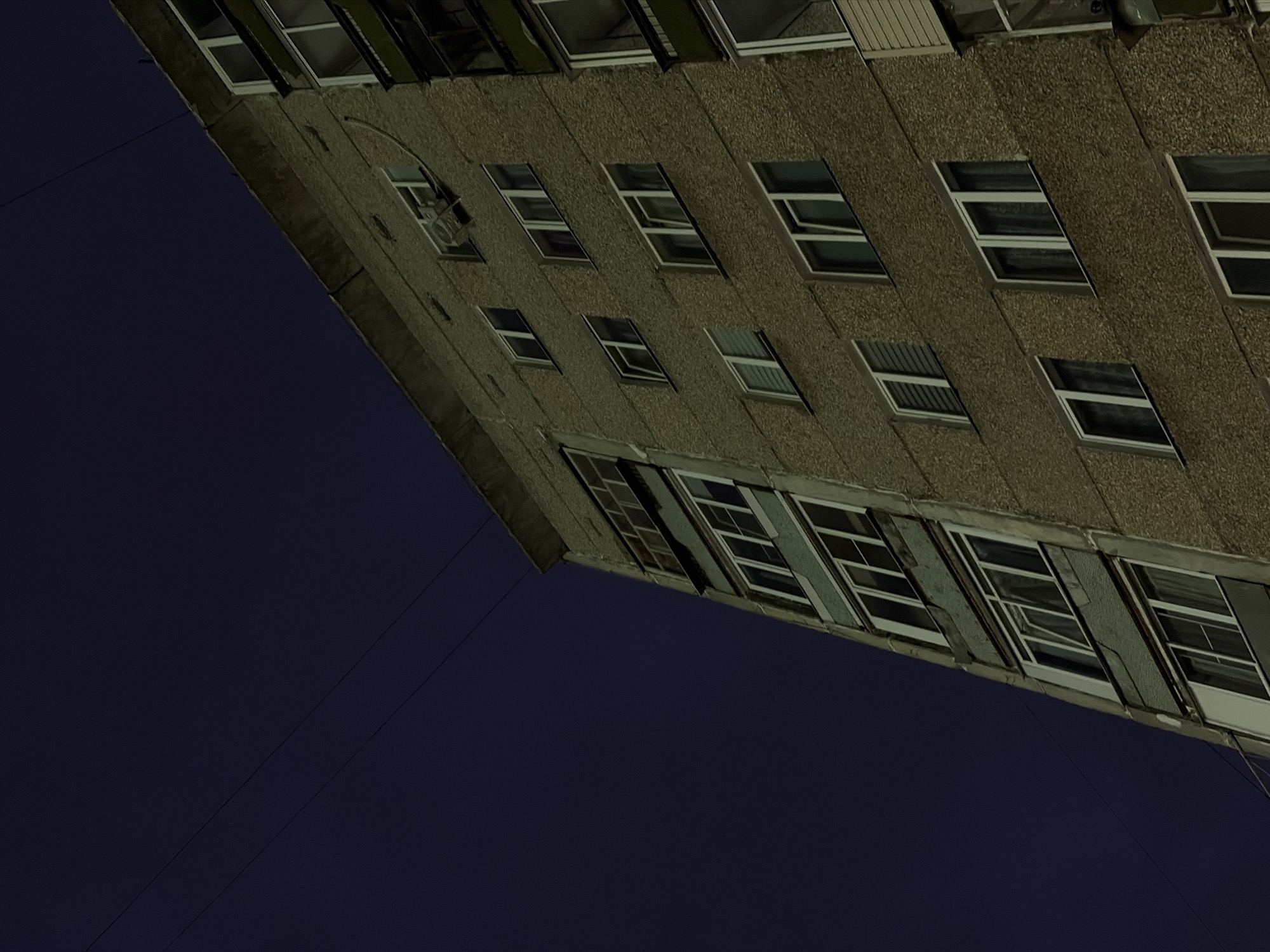 An exterior, low-angle, medium shot captures the side of a multi-story concrete apartment building against a dark night sky. The building's facade is textured and appears worn, with several windows visible. Some windows are open, revealing darker interiors, while others are closed and reflect the dim light. The building is angled to the right of the frame, with its roofline extending to the top right corner. A thin power line stretches diagonally across the dark sky from the upper left towards the building. No people are visible in the scene. The overall mood is somber and quiet, typical of a residential area at night.