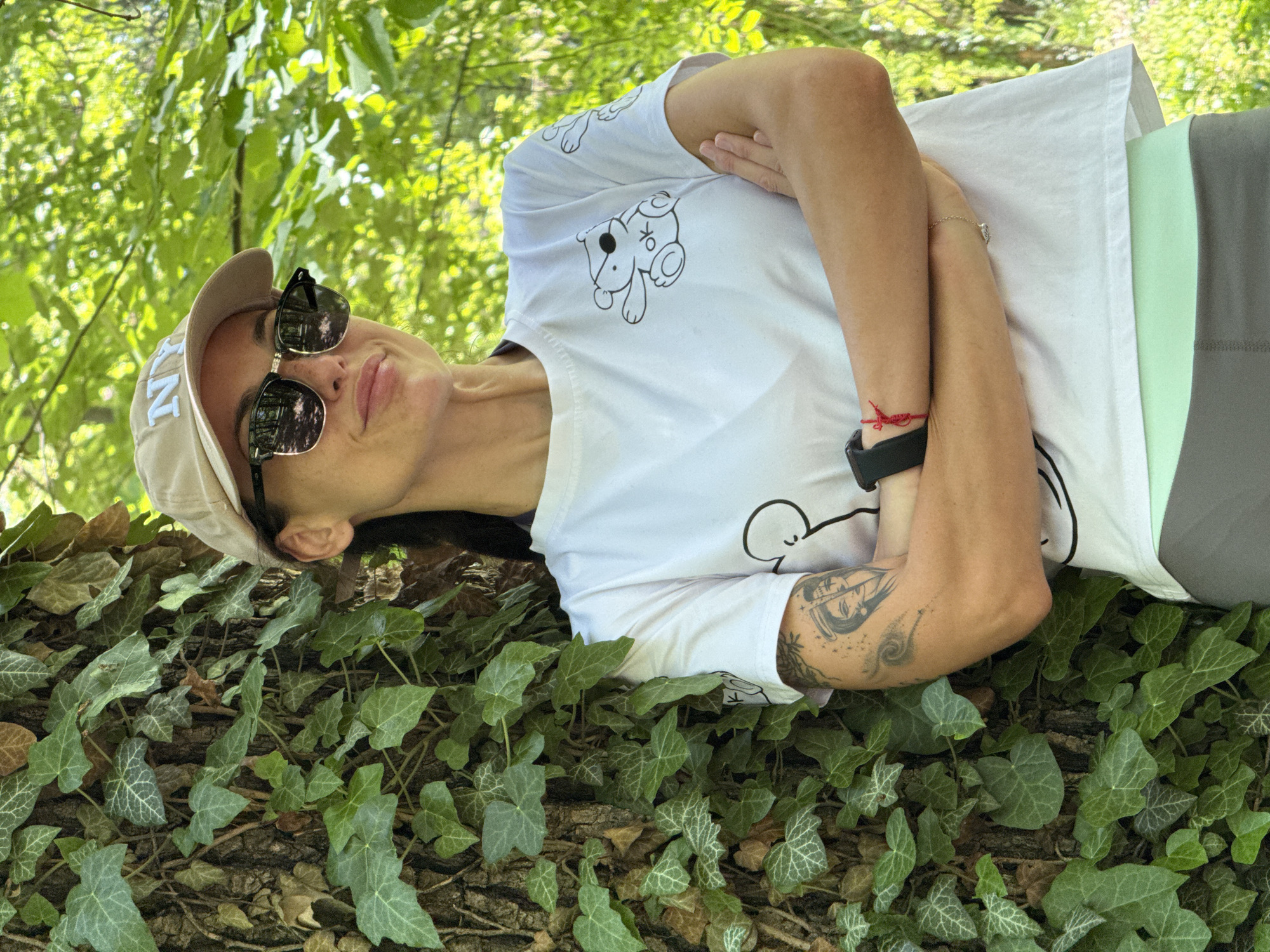 A woman lies on a tree trunk covered in ivy, under a canopy of green leaves. She is wearing a light-colored baseball cap, sunglasses, a white t-shirt with cartoon animal designs, and a black watch. Her arms are crossed over her chest. The photo was taken outdoors, seemingly in a park or forested area, but the location is not stated explicitly to be the city of Pleven, Bulgaria.
