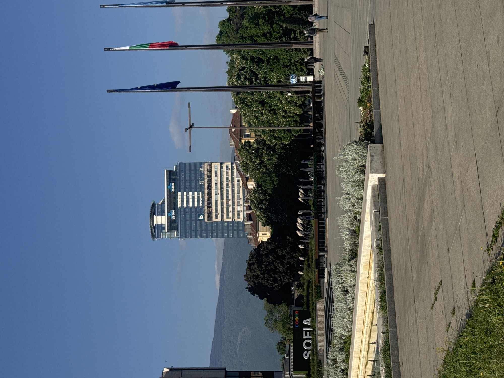 The image shows a view of Sofia, Bulgaria, with a prominent modern building featuring reflective glass facades under a clear blue sky. The Vitosha Mountain is visible in the background, creating a scenic backdrop. In the foreground, flagpoles fly several flags, including what appears to be the Bulgarian and European Union flags. Along the side, a pedestrian walkway with gray stone tiles borders landscaped areas. A sign displaying "SOFIA" is visible. People are walking in the distance.