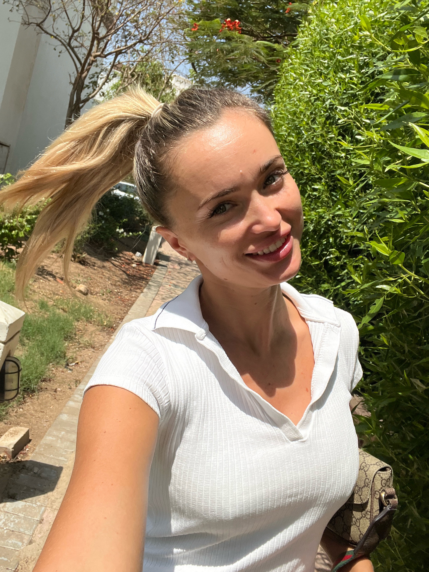 A woman with blonde hair in a high ponytail and green eyes smiles at the camera, holding a tan Gucci handbag. She is wearing a white, ribbed polo shirt and stands outdoors with a lush green hedge behind her and a paved walkway and some dry vegetation to her left. The scene appears to be in Sharm El Sheikh, Egypt, given the warm, sunny weather and the presence of palm trees in the background.