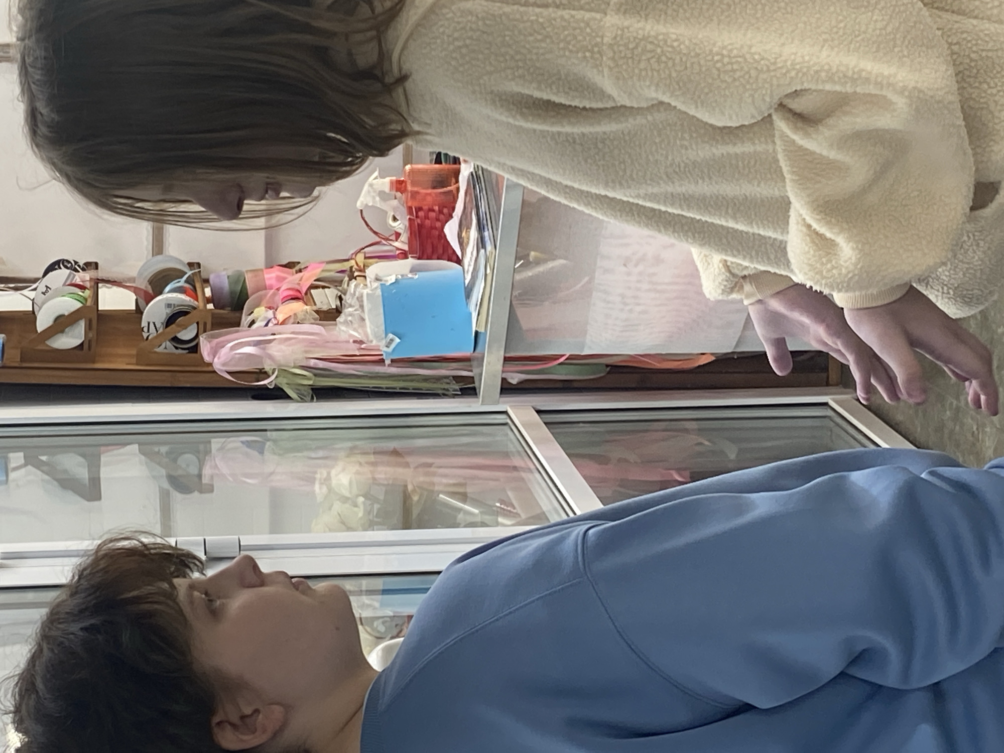 Two people stand on either side of a counter. A person with short dark hair, wearing a light blue shirt, stands on the bottom of the image. The second person with long brown hair and wearing a cream-colored fleece jacket is on the top. The counter is cluttered with art and craft supplies, including ribbons, containers, and a blue box. The bottom half of the counter is glass. The location is unknown and there is no way to determine if this picture was taken in Yekaterinburg, Russia.