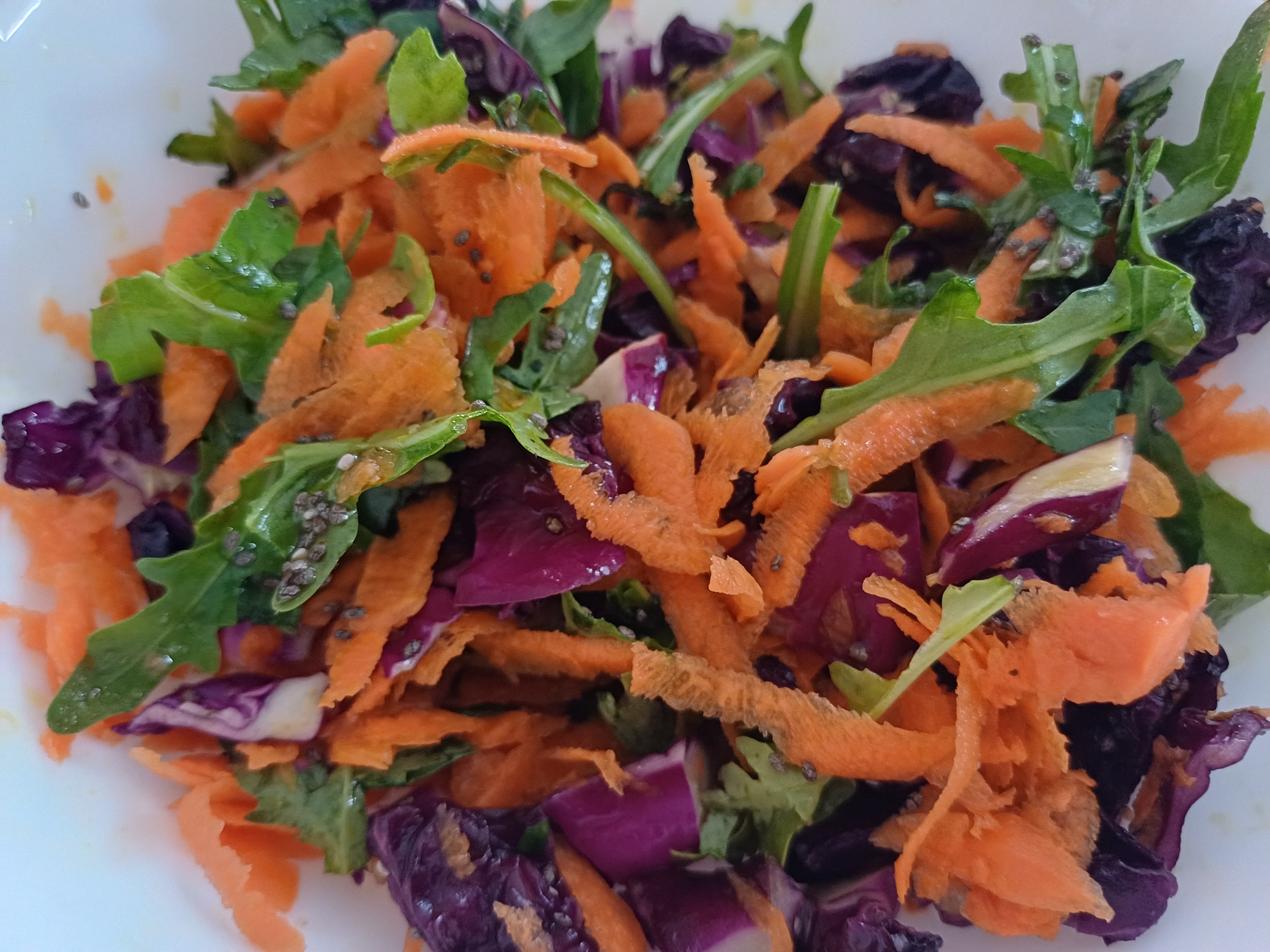 A close-up, overhead shot shows a colorful salad in a white bowl. The salad is composed of shredded carrots, dark purple cabbage, and arugula greens. Tiny black seeds, possibly chia seeds, are scattered throughout the salad. The lighting is bright and natural, suggesting the photo was taken indoors during the day. There is no indication of the location being in the city of Чёрновский, Russia.