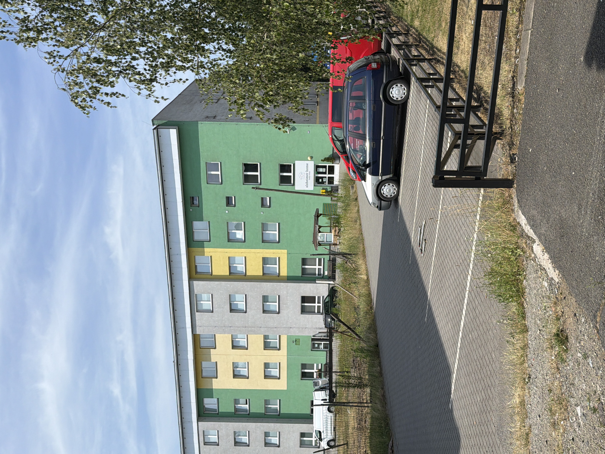 This image shows a three-story apartment building with a multi-colored facade in gray, yellow, and green. The building has numerous windows on each floor. In front of the building is a parking lot paved with interlocking bricks, with white parking lines. Several vehicles are parked in the lot, including a dark blue minivan, a red van, and some white vans closer to the building. A black metal fence separates the parking lot from a grassy area. There is a tree with green leaves in the foreground, partially obscuring the right side of the building. A sign on the building reads "Alzheimer Home" with a logo. The sky is blue with scattered clouds. The image appears to be taken on a sunny day. The specific location is the Alzheimer Home in Český Těšín, Czechia.