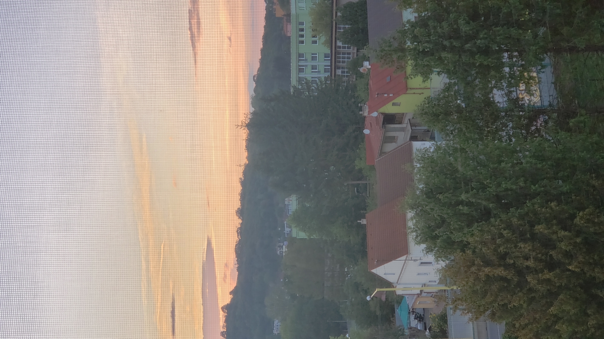 The image captures a view of a residential area in Prešov, Slovakia, likely during sunset or sunrise, as indicated by the warm hues in the sky. A fine mesh, possibly from a window screen, is visible in the foreground, overlaying the entire scene. The landscape features rolling hills covered in dense green foliage in the background.
In the mid-ground, several buildings are visible. There's a prominent light green building with multiple floors and numerous windows, characteristic of an apartment complex or a public facility. Adjacent to it, a cluster of houses with reddish-brown tile roofs are present, suggesting a suburban or urban neighborhood. Some of these houses have visible architectural details like balconies and chimneys. Trees, varying in size and density, are scattered throughout the scene, some in front of the buildings and others integrated into the landscape.
The foreground is dominated by the tops of trees, their leaves creating a textured green canopy. A yellow pole, possibly part of a lamppost or street light, is visible on the left side of the frame, near a small building with a green umbrella and some seating outside. The overall impression is a tranquil, everyday scene within a European city, viewed through a window or screen. There are no people or animals clearly depicted in the image.