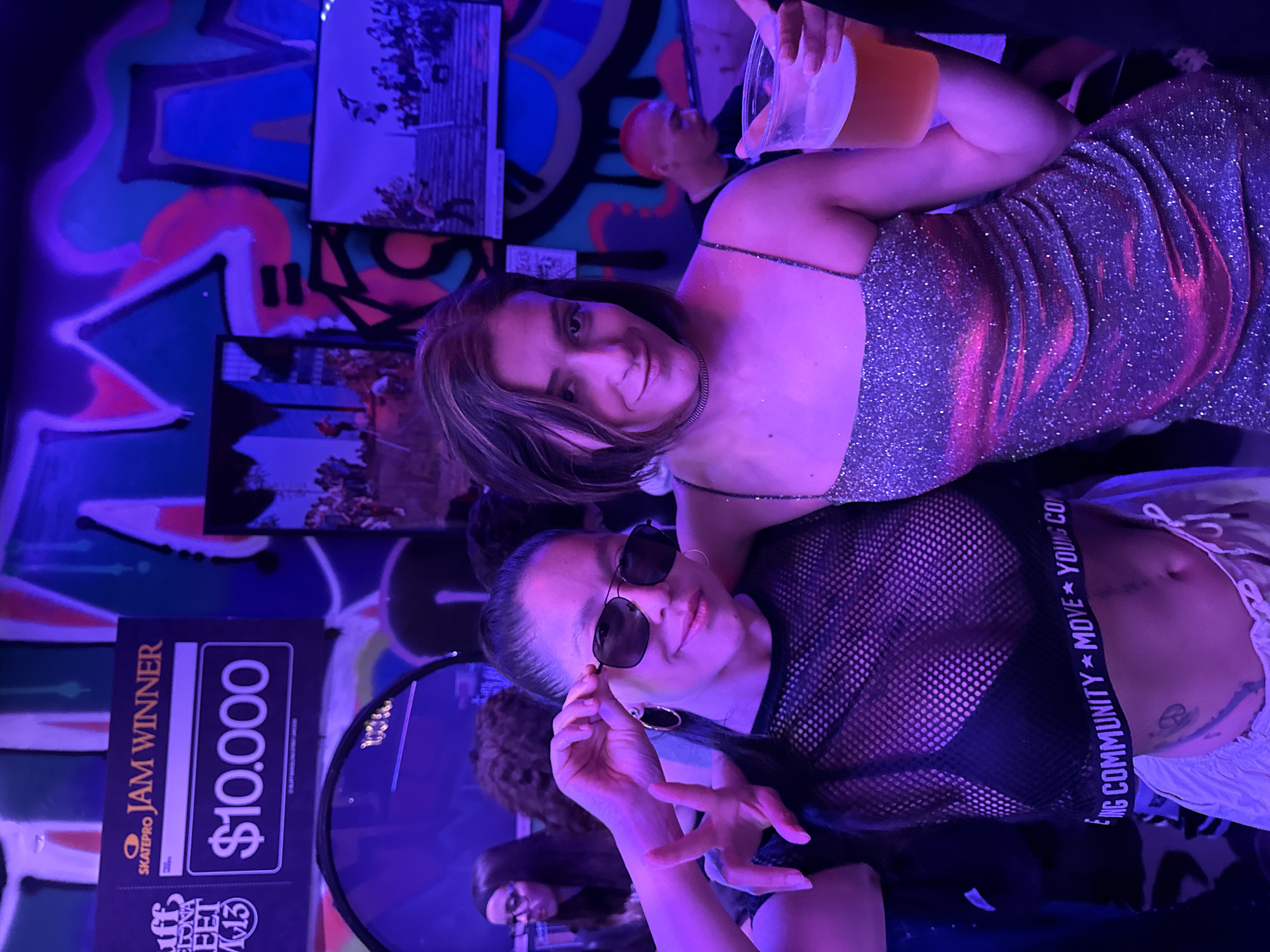 Two young women pose for a photo indoors against a background of graffiti and event-related signage. The woman in the foreground wears sunglasses, a black mesh top, and white shorts. A black band with white lettering "ING COMMUNITY * MOVE * YOUNG CO" is visible on her lower torso. The woman behind her wears a glittery dress and holds a drink in a plastic cup. A man with a shaved red head stands in the background. A sign to the left indicates "SKATEPRO JAM WINNER $10.000" and mentions "SUFF BARCELONA STREET M13." The environment is lit with bright blue and purple lights. The location is Barcelona, Spain, as indicated on the signage.