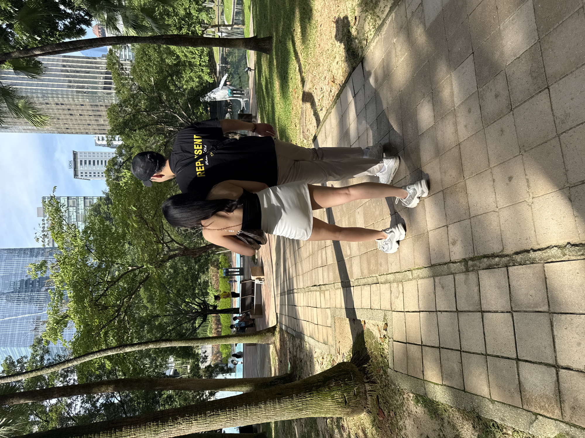 The media file shows two individuals, a man and a woman, strolling away from the viewer on a paved pathway within an urban park in Kuala Lumpur, Malaysia. The man, wearing a black cap, a black t-shirt with yellow text "REP SENT Fashion Club", and khaki pants, walks beside the woman. She is dressed in a black top, a white mini-skirt, and white chunky sneakers, carrying a small dark handbag with a chain strap.
Strong shadows from trees and the individuals fall across the square-paver pathway, indicating a sunny day, likely mid-morning or afternoon. The weather appears clear and bright. The park features lush green trees and grassy areas. In the background, several modern high-rise buildings are visible, with one distinctive glass-facade structure prominently featuring the "UOA" logo. Further down the path, other people can be observed sitting on a park bench and near a water feature. The scene depicts a serene and sunny walk amidst urban greenery.
