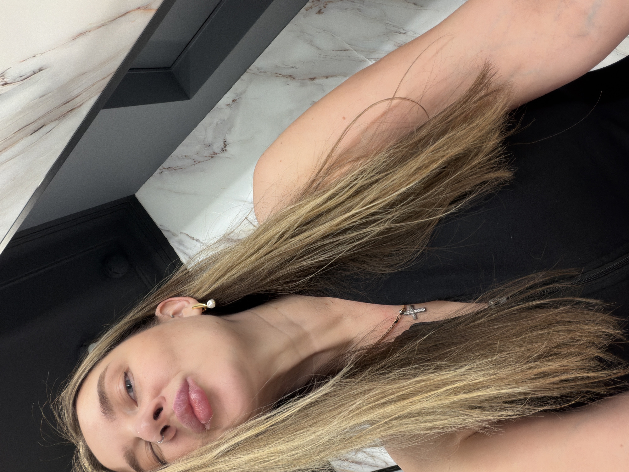 A selfie-style image depicts a young woman with long, blonde hair. She is wearing a black top and a gold earring with a pearl accent. A gold necklace with a cross pendant is visible on her neck. The background consists of a black surface and a marble-patterned wall. The location is not discernible from the image.
