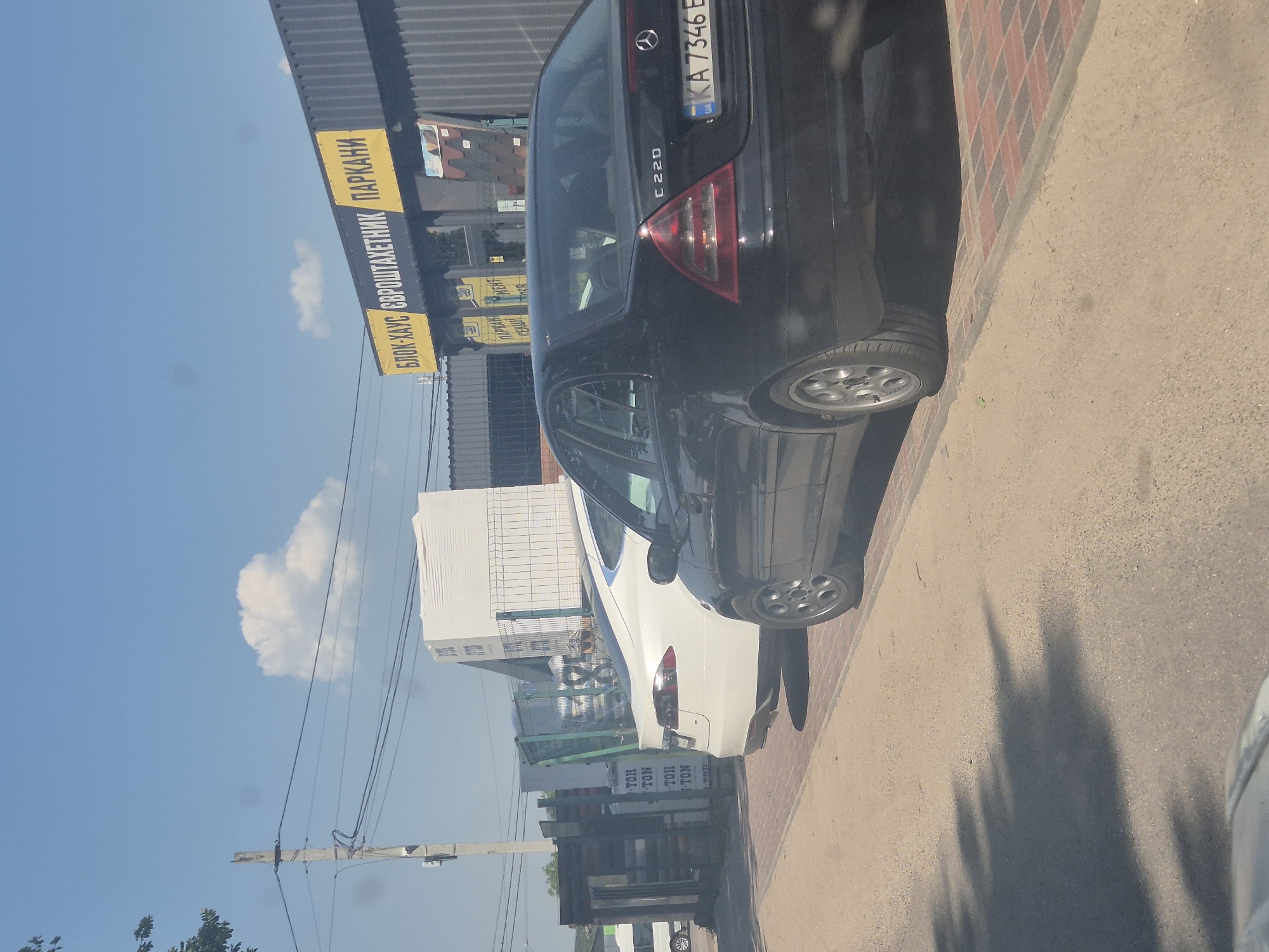 The image shows a street scene with two cars, a white Tesla and a black Mercedes-Benz, parked on a brick-patterned sidewalk next to a dirt road. In the background, there is a building with a sign in Ukrainian that reads "БЛОК-ХАУС ЄВРОШТАКЕТНИК ПАРКАНИ". There are also stacks of white building materials, possibly insulation panels, on pallets behind a green fence. The sky is blue with a few scattered clouds, and the sun is casting shadows on the ground. The overall impression is a commercial area, possibly a building materials supplier, on a sunny day in Pidhaitsi, Ukraine.
