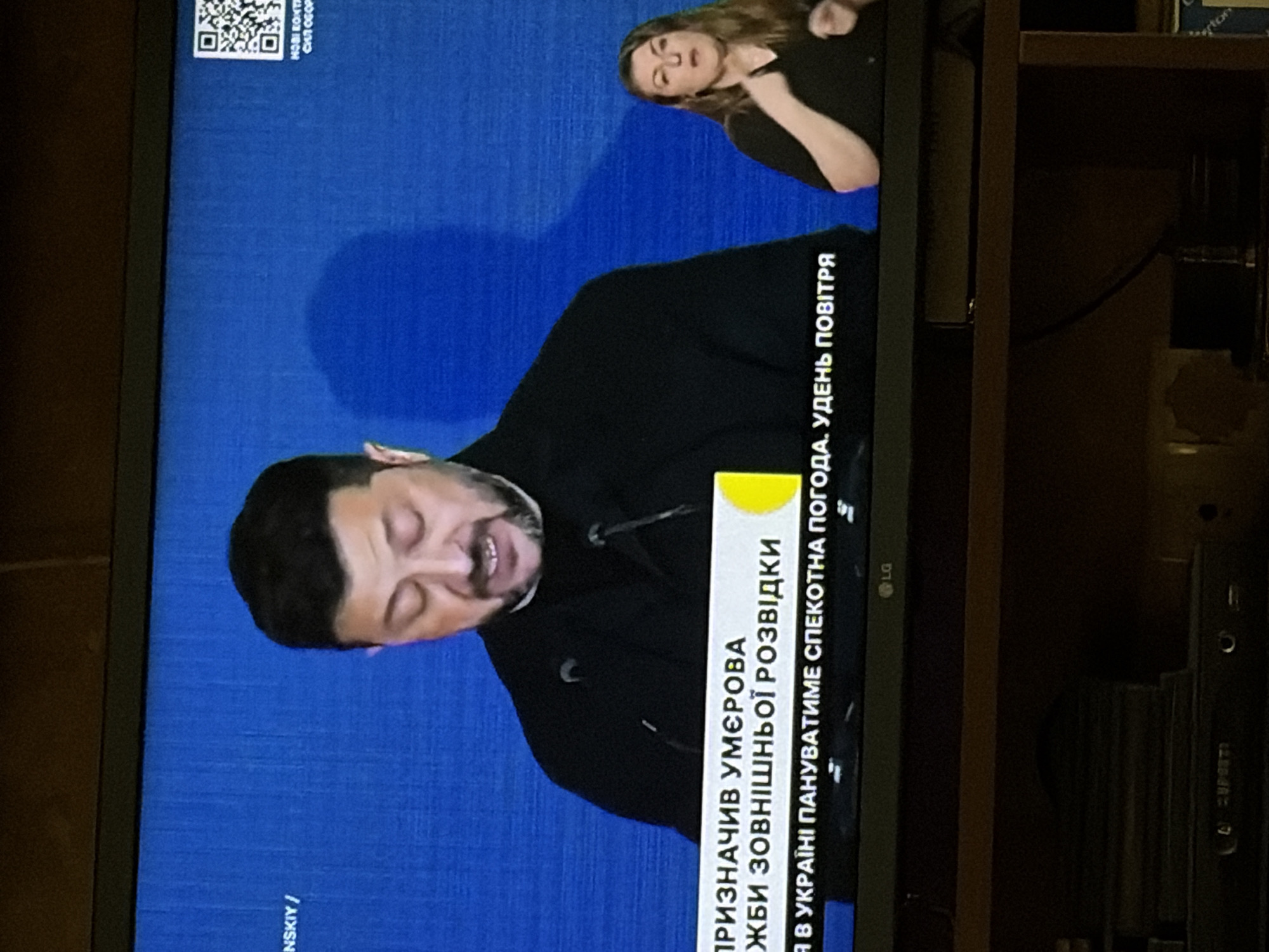 A television screen displays a man and a woman. The man, who appears to be speaking, is in the center of the screen. He is wearing a dark, collared shirt and has short, dark hair and a beard. His eyes are closed and his mouth is slightly open. To the right of the man, a woman is visible, facing left. She has long, brown hair and is wearing a black top. Her hands are raised and appear to be gesturing. A blue background is behind the people, with some text and a QR code in the upper right corner. The bottom of the screen shows a white banner with Ukrainian text. Below the screen, a dark wooden surface is visible, with some electronic equipment stacked on it. The lighting appears to be from a room, reflecting slightly off the screen.