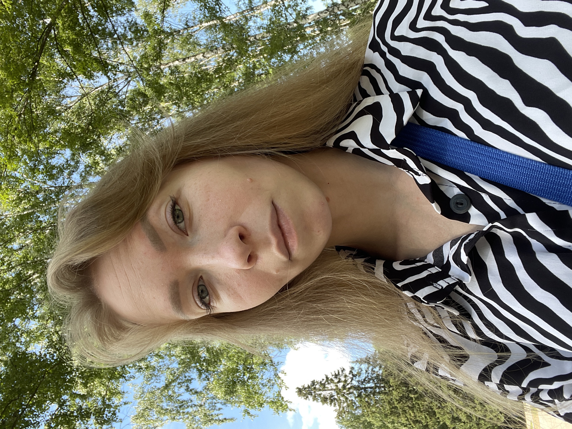 A young woman with long blonde hair is taking a selfie outdoors. She is wearing a black and white zebra-print shirt and a blue strap is visible across her chest. The background shows green trees and a bright blue sky with some clouds. The image appears to have been taken in a park or natural setting, and the lighting suggests it is daytime. There is no information in the image that would indicate a specific location in the city of Perm, Russia.