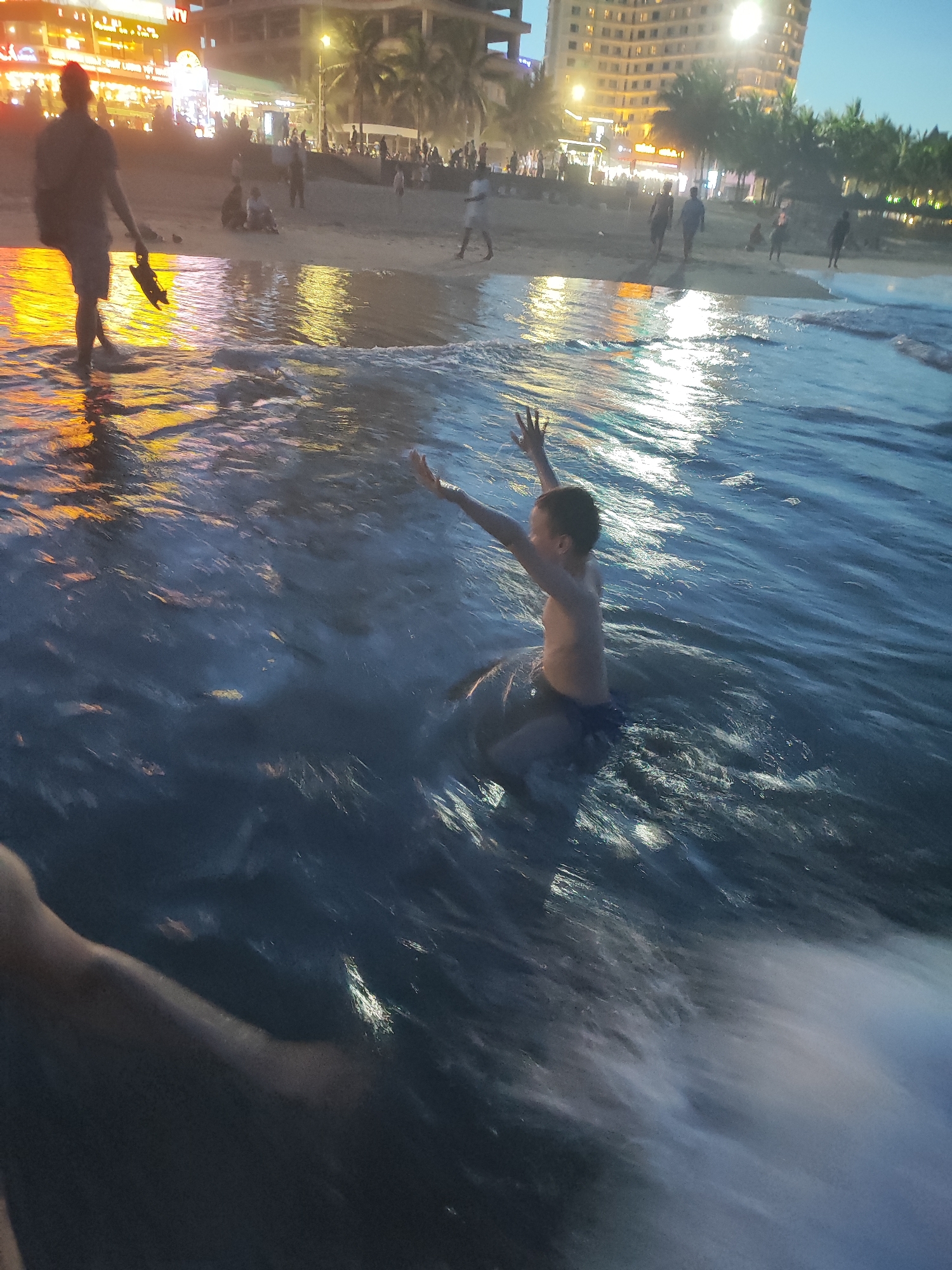 A wide-angle shot captures a nighttime scene on a beach in Đà Nẵng, Vietnam. A young boy sits in the water with arms raised, appearing to be enjoying the waves. Further back, people stroll along the shore near buildings lit up with bright lights. To the left, a person wades into the water carrying footwear. Palm trees are visible along the coastline. The water is choppy, reflecting the ambient light. The foreground includes a partial view of an arm.