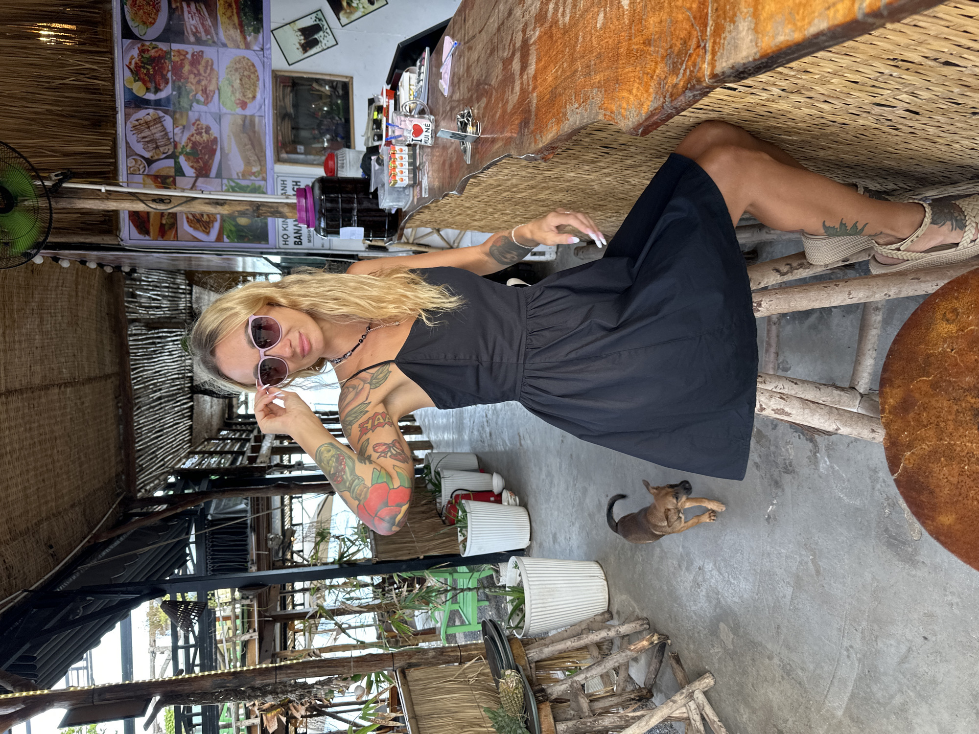 A blonde woman wearing sunglasses and a black dress sits on a stool at a rustic bar in a beachfront establishment in Phu Thuy Ward, Vietnam. She is posing with one hand touching her sunglasses. A small brown dog walks on the concrete floor to her left. The bar is made of wood and woven material, with various items displayed on it, including bottles and a mug. A menu with food pictures is displayed on the wall behind the bar. The environment appears to be outdoors, with natural lighting and vegetation visible in the background.