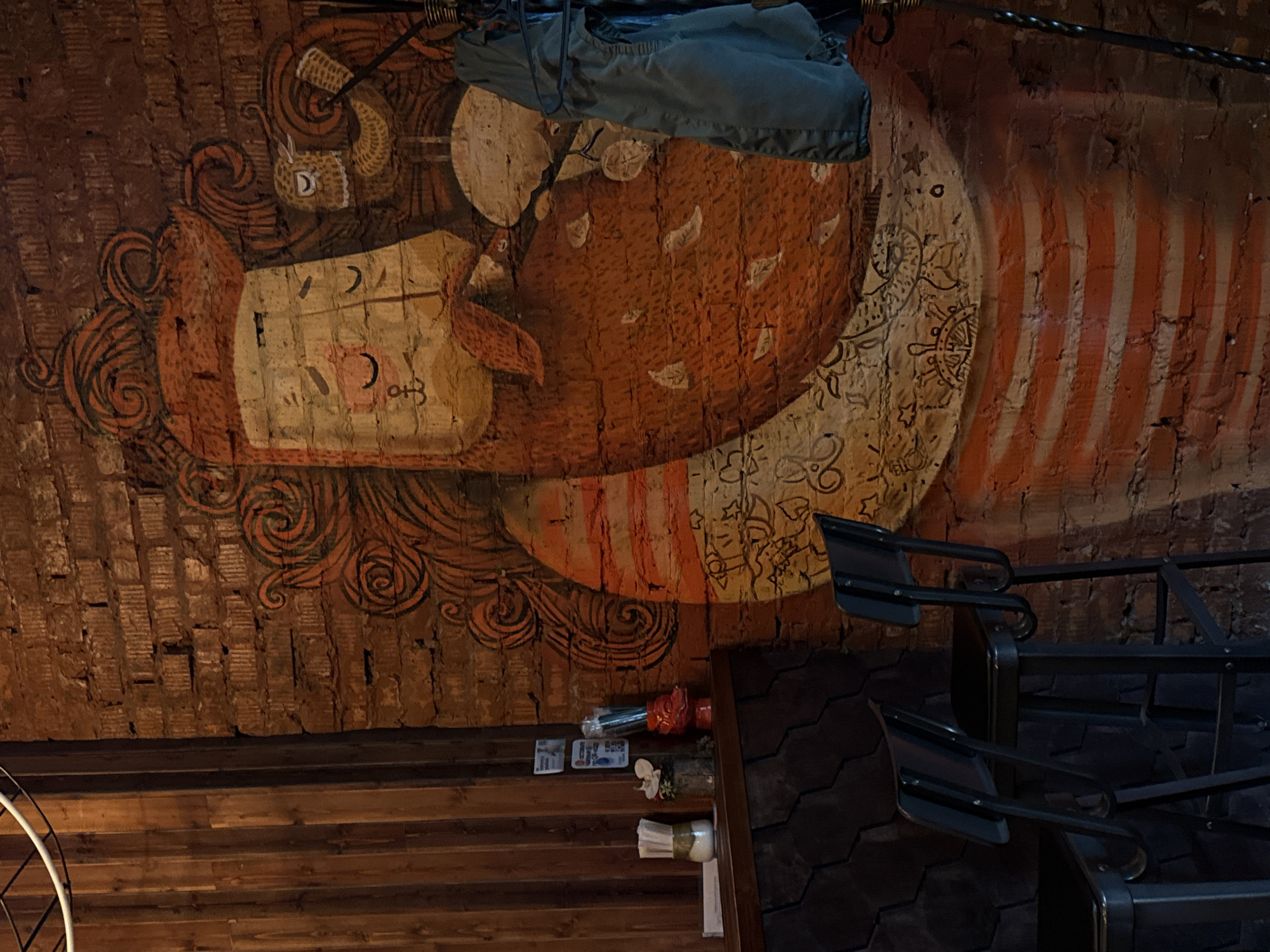 The image, rotated 90 degrees clockwise, depicts an interior scene, likely within a cafe, restaurant, or bar in Birobidzhan, Russia. A large, intricate mural is painted directly onto an exposed brick wall or ceiling.
The central subject of the mural is a stylized, cartoon-like face, oriented upright if the image were rotated counter-clockwise. The face has closed eyes, a prominent beard or mane rendered in flowing, orange-brown swirling patterns, and light-colored cheek areas adorned with crescent moon shapes and other symbols. Perched above its head is a small, light-colored creature, possibly a squirrel or similar rodent, with a patterned tail, holding a small object. Behind the face is a large, rounded, reddish-brown shape featuring scattered white, leaf-like motifs. To the right of the central figure, the mural extends with a section containing delicate black line drawings, depicting stars, ship wheels, and floral patterns on a light background, transitioning into bold, vertical reddish-orange stripes on the far right. The entire mural is characterized by its warm color palette and swirling, organic designs.
Below the mural (and to the left in the current orientation) are horizontal wooden beams or paneling. In the lower right corner, dark, textured walling (possibly tiles or stone) is visible, along with black metal frames, possibly parts of chairs or decorative fixtures. On a wooden ledge or shelf, several small items are arranged: a red container holding what appears to be cutlery or straws, a couple of small white cards with illegible text, a white flower in a small vase, and a bundle of white napkins tied with a light-colored ribbon. A dark blue or grey piece of fabric or clothing hangs from a rod in the upper part of the image, partially obscuring the mural. The scene is illuminated by artificial, warm lighting, contributing to a cozy and artistic ambiance. No specific activity is depicted.