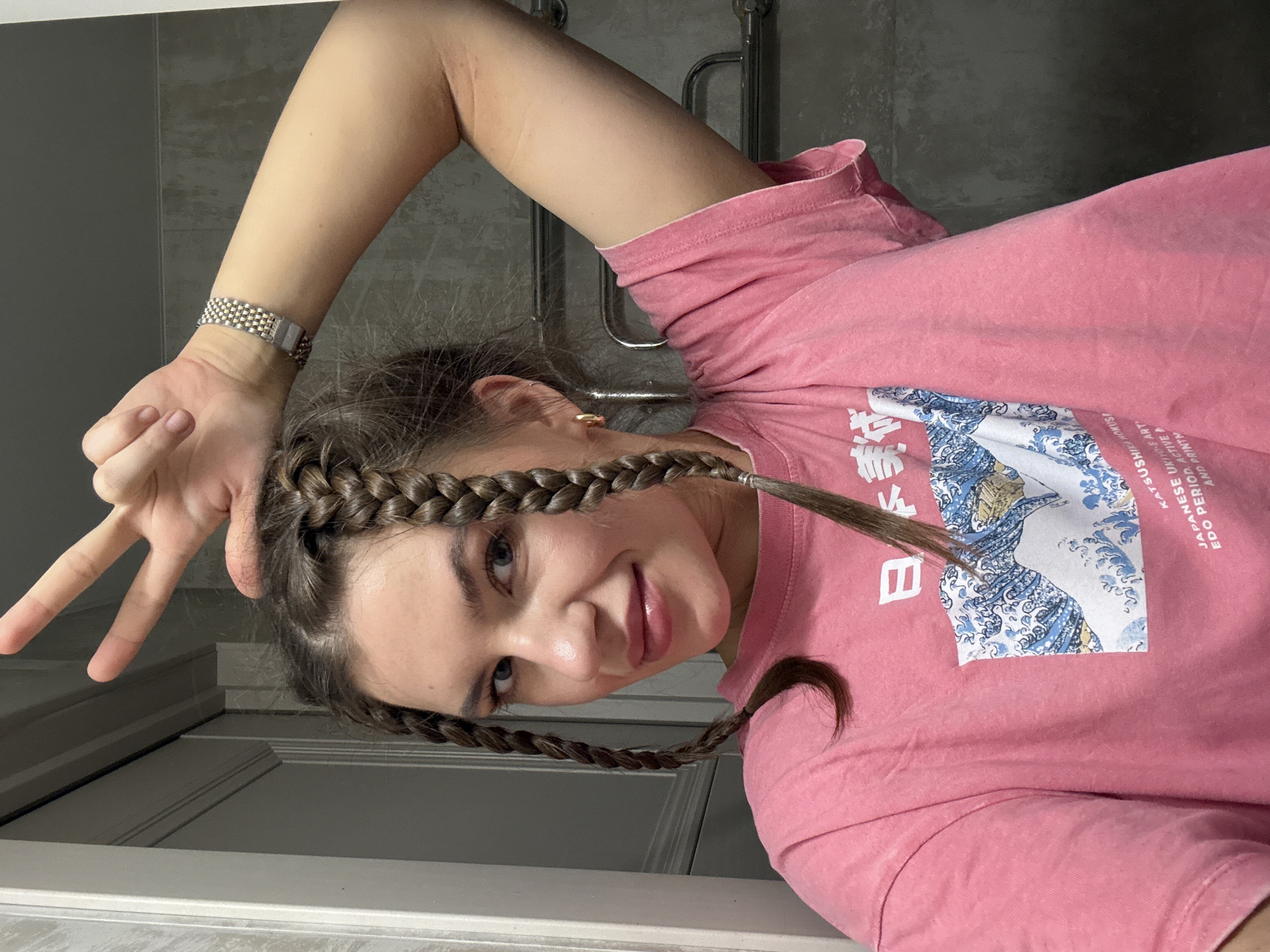 A woman with brown braided hair is indoors, wearing a pink t-shirt with Japanese art printed on it and a silver wristwatch. She is holding her hand up making a peace sign. The background includes a gray tiled wall and a white door.