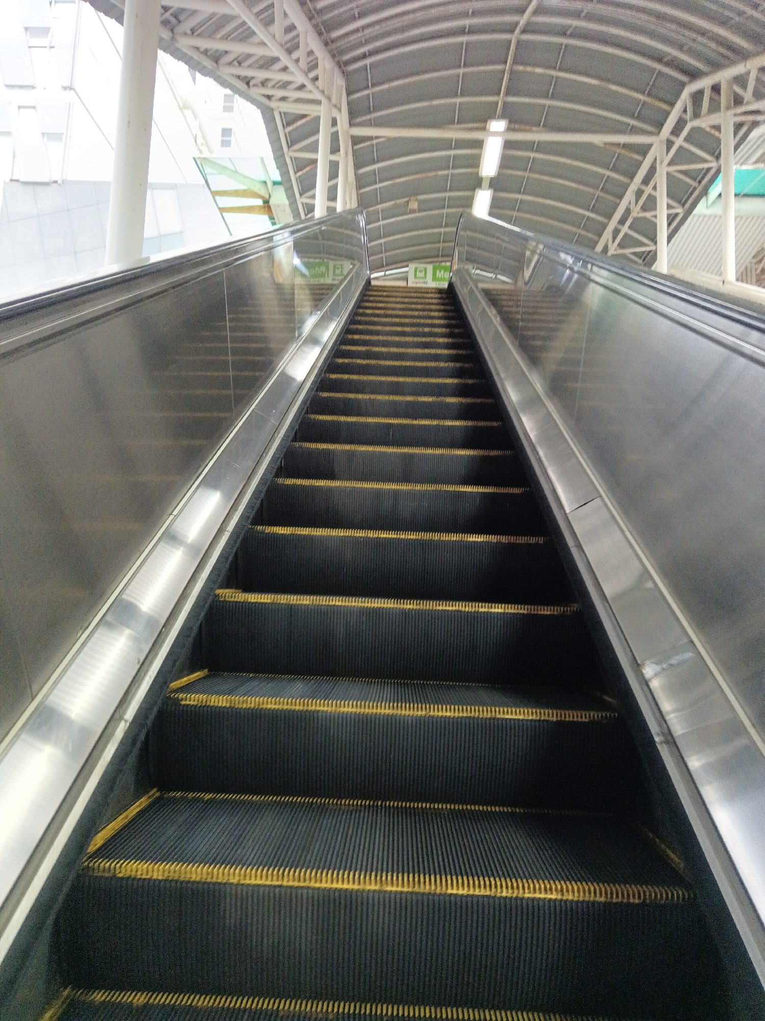 An upward-facing shot of an empty escalator shows its dark treads with yellow warning stripes, set against a metallic handrail and a silver side panel. Above, a curved metal roof structure with white support beams and fluorescent lighting arches over the escalator. In the background, a modern building with glass windows is visible, suggesting an urban setting. The overall impression is that of a public transportation hub or a large building. The sign visible on the left side of the escalator reads "Masjid Jamek" which is a landmark in Kuala Lumpur, Malaysia.