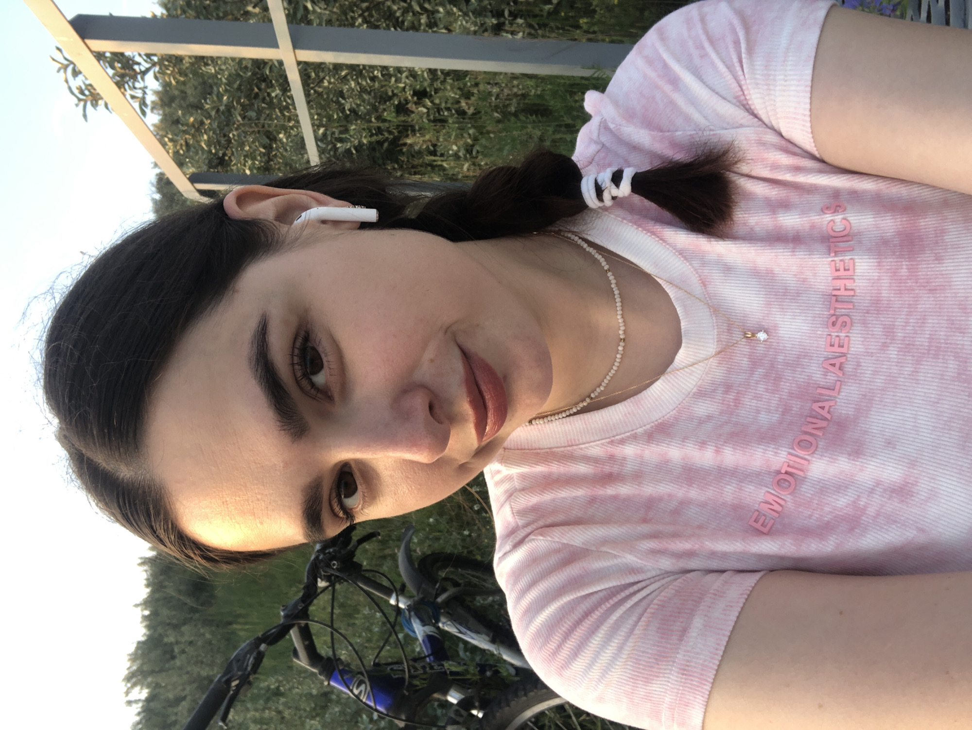 A young woman with dark hair in a braid is taking a selfie outdoors. She is wearing a pink tie-dye t-shirt with "EMOTIONAL AESTHETICS" printed on it, a pearl necklace, and a gold pendant. She has white earbuds in her ears. A bicycle is visible behind her to the left, and a metal fence and trees are in the background. The setting appears to be a park or outdoor recreational area in Chaikovsky, Russia.
