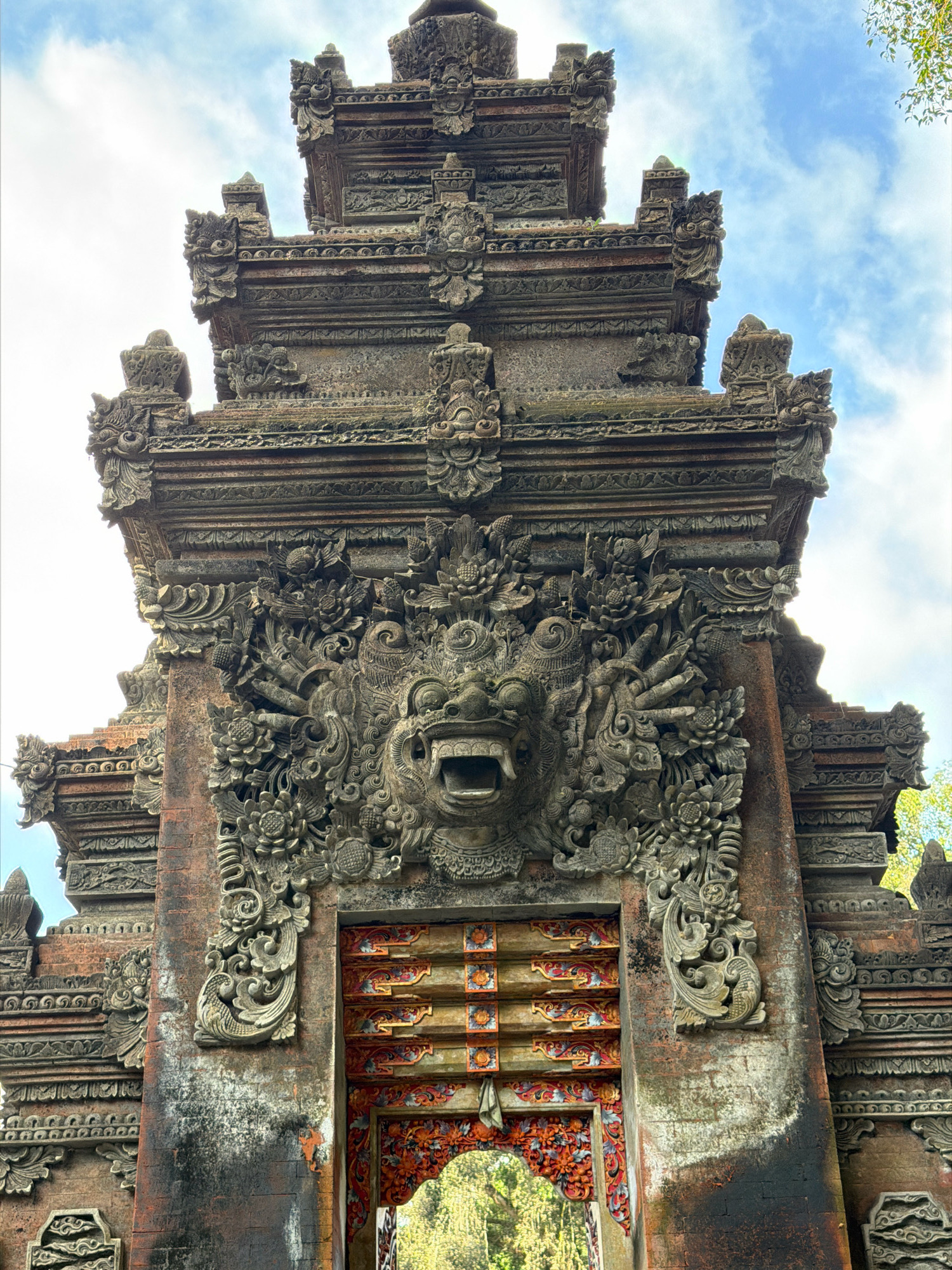The image shows a low-angle, close-up view of an ornate stone temple gate in Tampaksiring, Indonesia. The gate is intricately carved with traditional Balinese motifs. Dominating the center is a fearsome demon's face with a gaping mouth and sharp teeth, surrounded by floral and serpentine elements. The stonework appears weathered and aged, with some areas showing patches of moss and discoloration. Above the demon's face, multiple tiers of layered stonework form a pagoda-like structure, also adorned with detailed carvings. The entrance itself is visible at the bottom, framed by decorative elements and painted with vibrant colors in red, orange, and gold. The sky, partially visible at the top, is blue with scattered white clouds, and the upper right corner shows some green foliage. The overall impression is one of ancient artistry and spiritual significance.