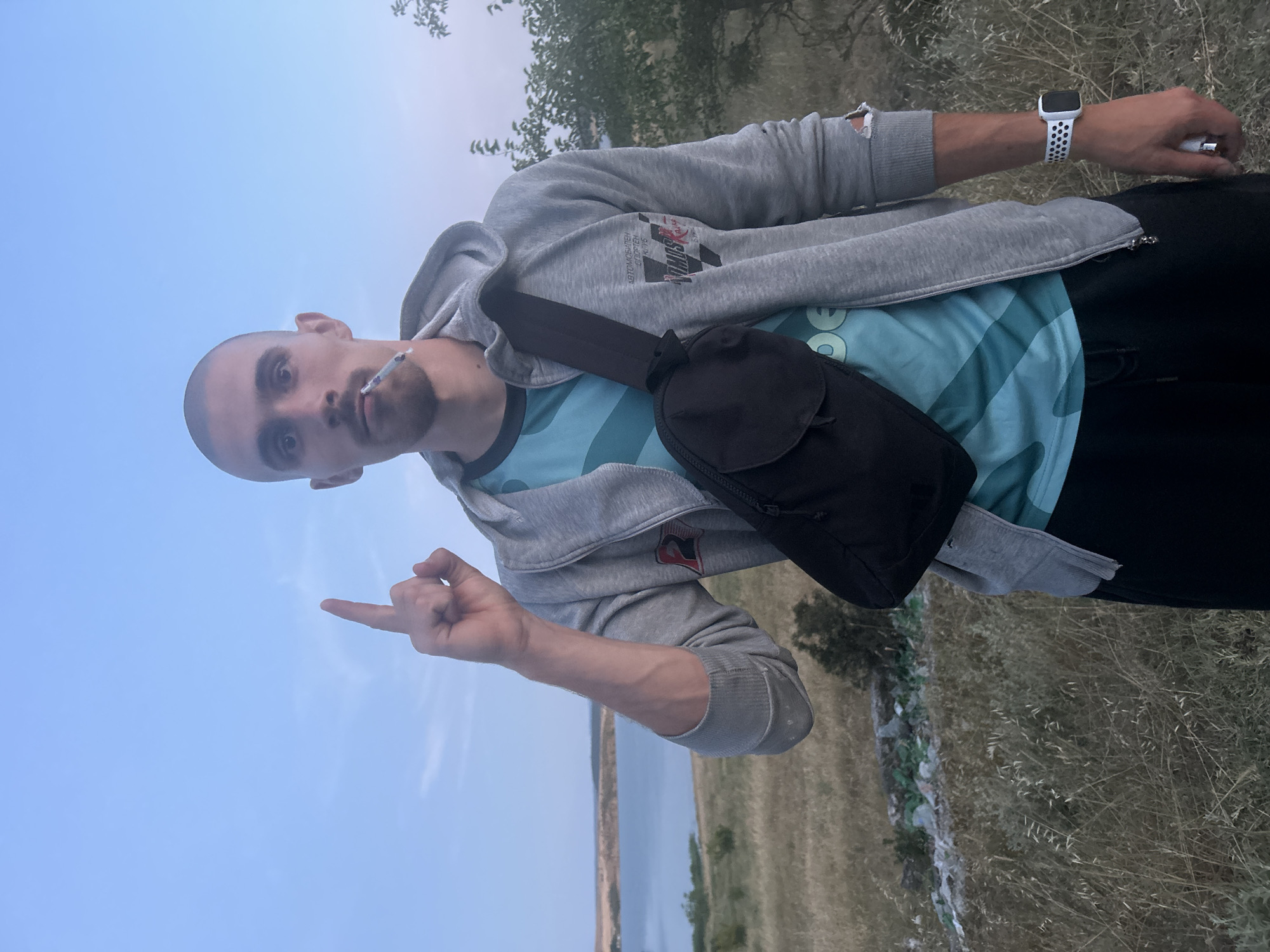 A young man stands outdoors with a cigarette in his mouth, pointing his index finger upwards. He is wearing a grey hooded jacket over a teal t-shirt with a geometric pattern, black pants, and a black crossbody bag. His left arm has a smartwatch with a white and black patterned band. The background shows a dry, grassy landscape with a body of water and a tree on the right. The sky is a pale blue with faint clouds. The man's pose and the presence of the cigarette suggest a moment of contemplation or emphasis. The environment appears to be a natural, possibly rural or undeveloped area, inconsistent with the city of Burgas, Bulgaria.