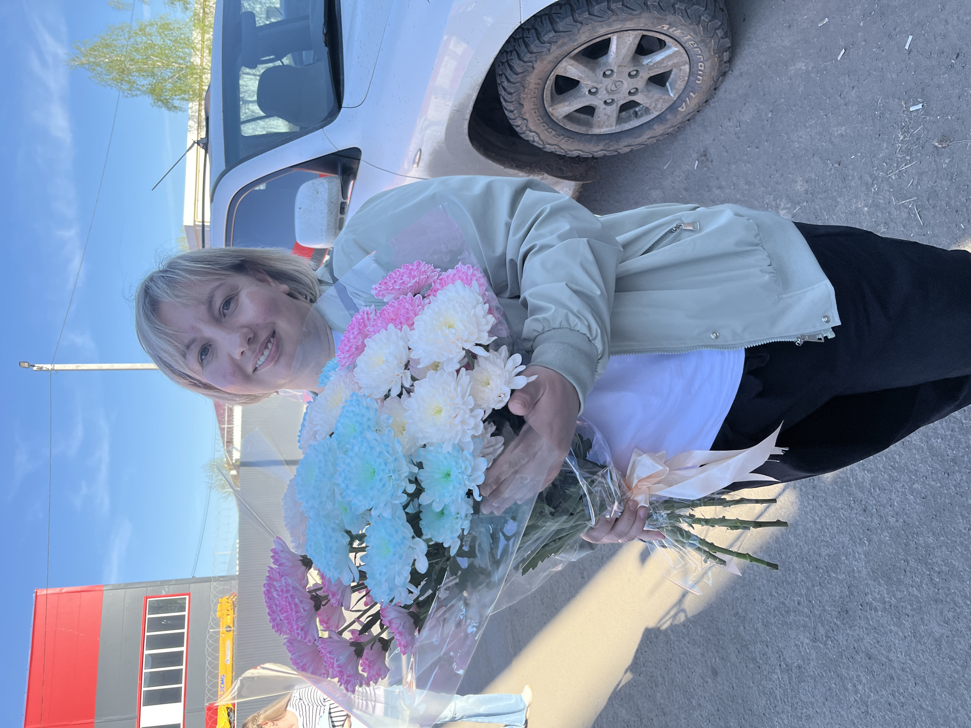 A woman with blonde hair smiles while holding a bouquet of pink, white, and blue flowers. She wears a light green jacket and black pants. A white car is partially visible in the background with a muddy tire. The background also shows a building with a red and gray facade under a blue sky. The photo was taken in Izhevsk, Russia.