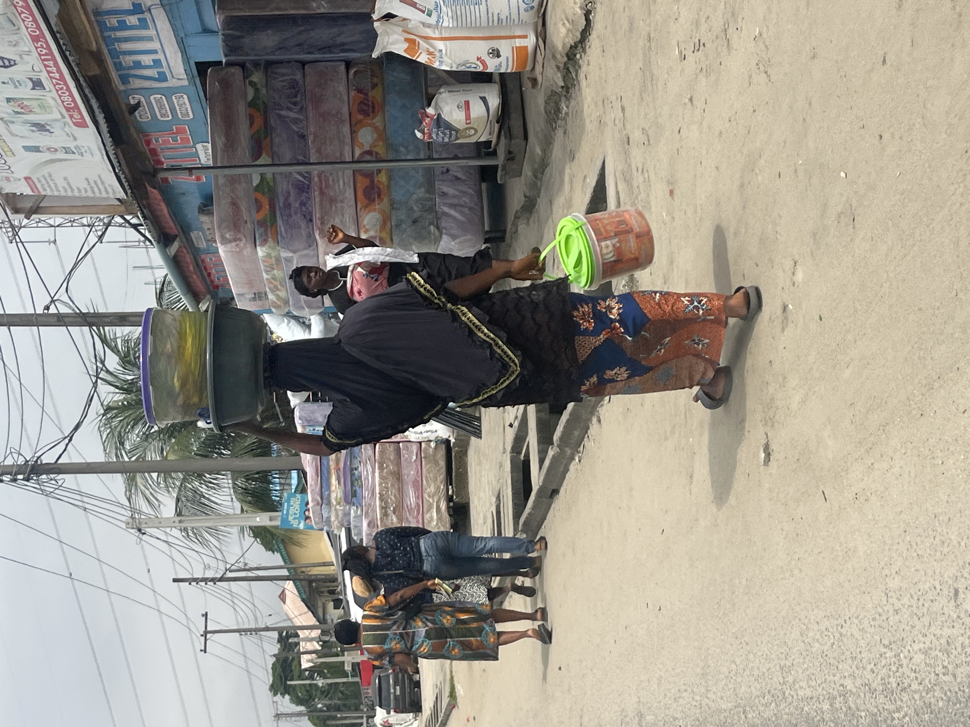 In Ado, Nigeria, a woman carrying a stack of round plastic containers and bowls on her head walks on a dusty road. She wears a black headscarf and a patterned orange and blue skirt. In her right hand, she carries a green plastic bucket filled with small items. To her left, two other women walk away from the camera, one in a colorful patterned dress and the other in blue jeans. Behind them, stacked mattresses are visible in front of a building with signs advertising "ZETTEL Interior." Power lines and poles are visible in the background under a cloudy sky. A black SUV is parked on the left side of the road. The scene depicts a typical street market environment.