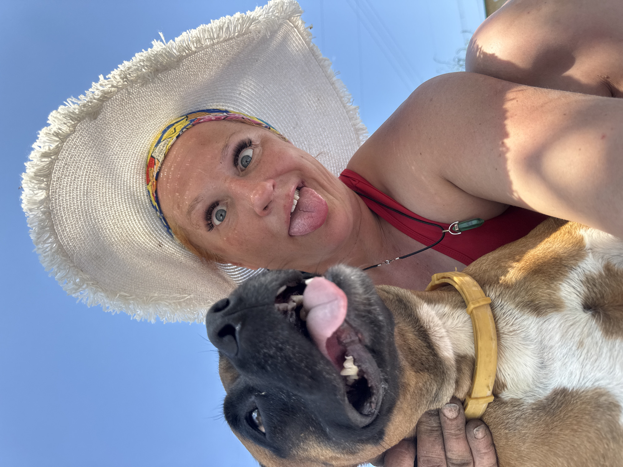 A woman and a dog are in the foreground of a low-angle shot against a bright blue sky. The woman, wearing a wide-brimmed straw hat with a colorful band, is looking directly at the camera with a playful expression, sticking out her tongue and crossing her eyes slightly. She is wearing a red swimsuit and a necklace. The dog, a fawn and white boxer, is directly below her and also looking at the camera with its tongue hanging out. The dog has a yellow collar. In the background is a clear blue sky, which suggests the photo was taken outdoors. The location is likely Chaikovsky, Russia.