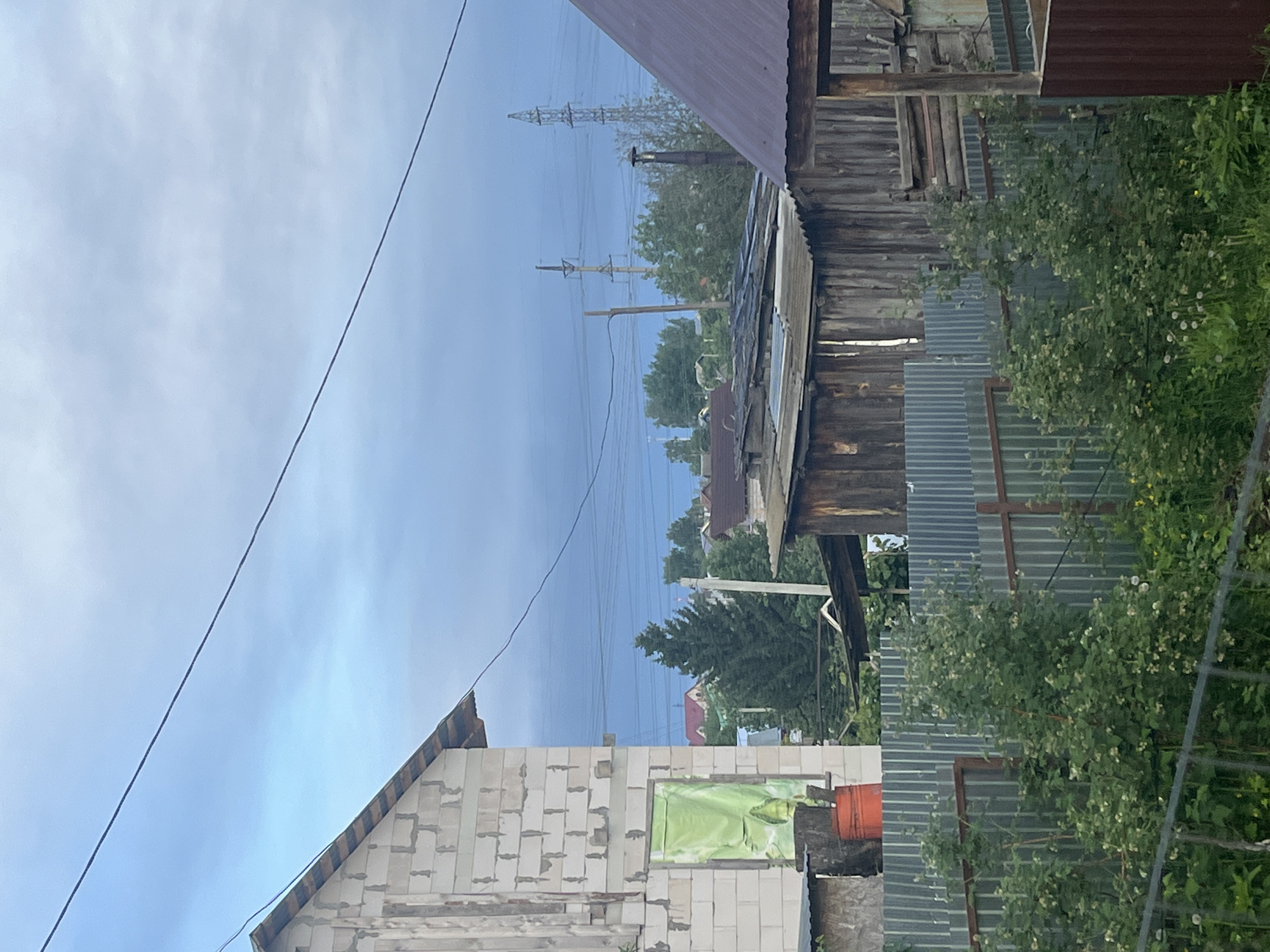 This outdoor scene shows a residential area in Chaikovsky, Russia. A partially constructed brick building is visible in the foreground. An orange bucket sits beside a window covered in green material. A corrugated metal fence runs along the property line, partially obscured by leafy bushes. Behind the fence stands an older, dilapidated wooden structure with a rusty roof. Overhead power lines traverse the sky, connected to utility poles. Trees dot the background, obscuring other houses and buildings. The sky is overcast and pale blue.