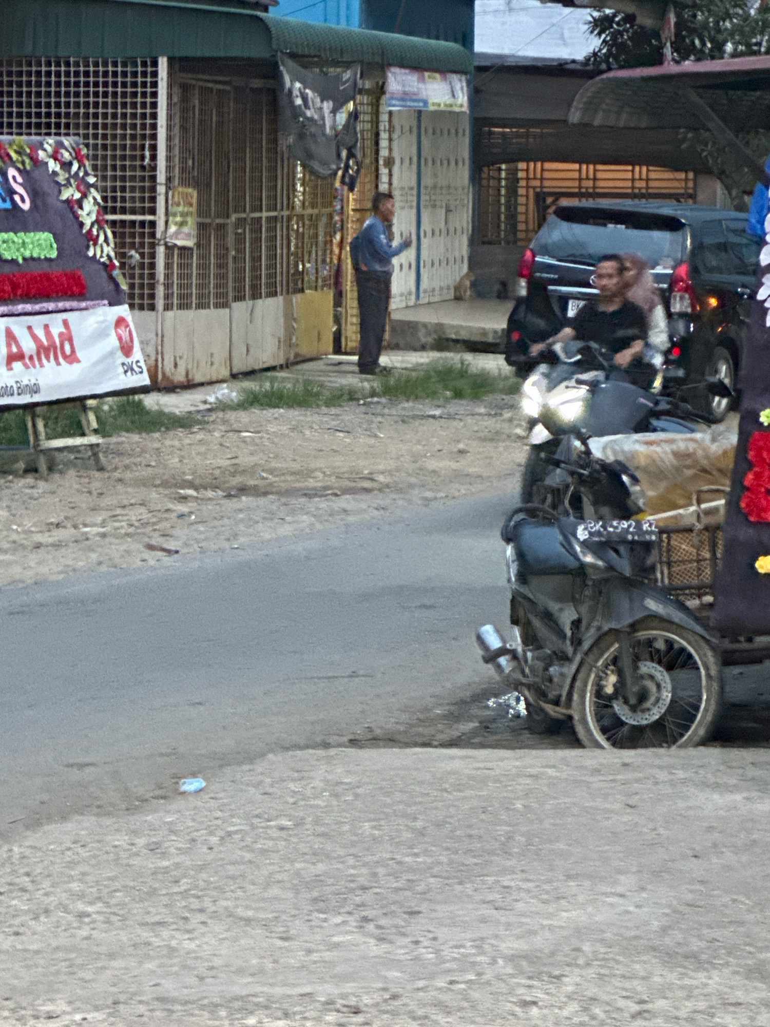 The image depicts a street scene in the city of Binjai, Indonesia, with a focus on the foreground and midground. A dark-colored motorcycle with a license plate reading "BK 592 RZ" is parked on the right side of the road. Behind the motorcycle, a black SUV is visible. In the midground, a man in a blue shirt and dark pants stands near a gated storefront. To the left, a banner with text, including "S", "A.Md", and "PKS", is displayed. The environment appears to be an urban setting with a paved road, a sidewalk, and several buildings in the background. The lighting suggests it is daytime.