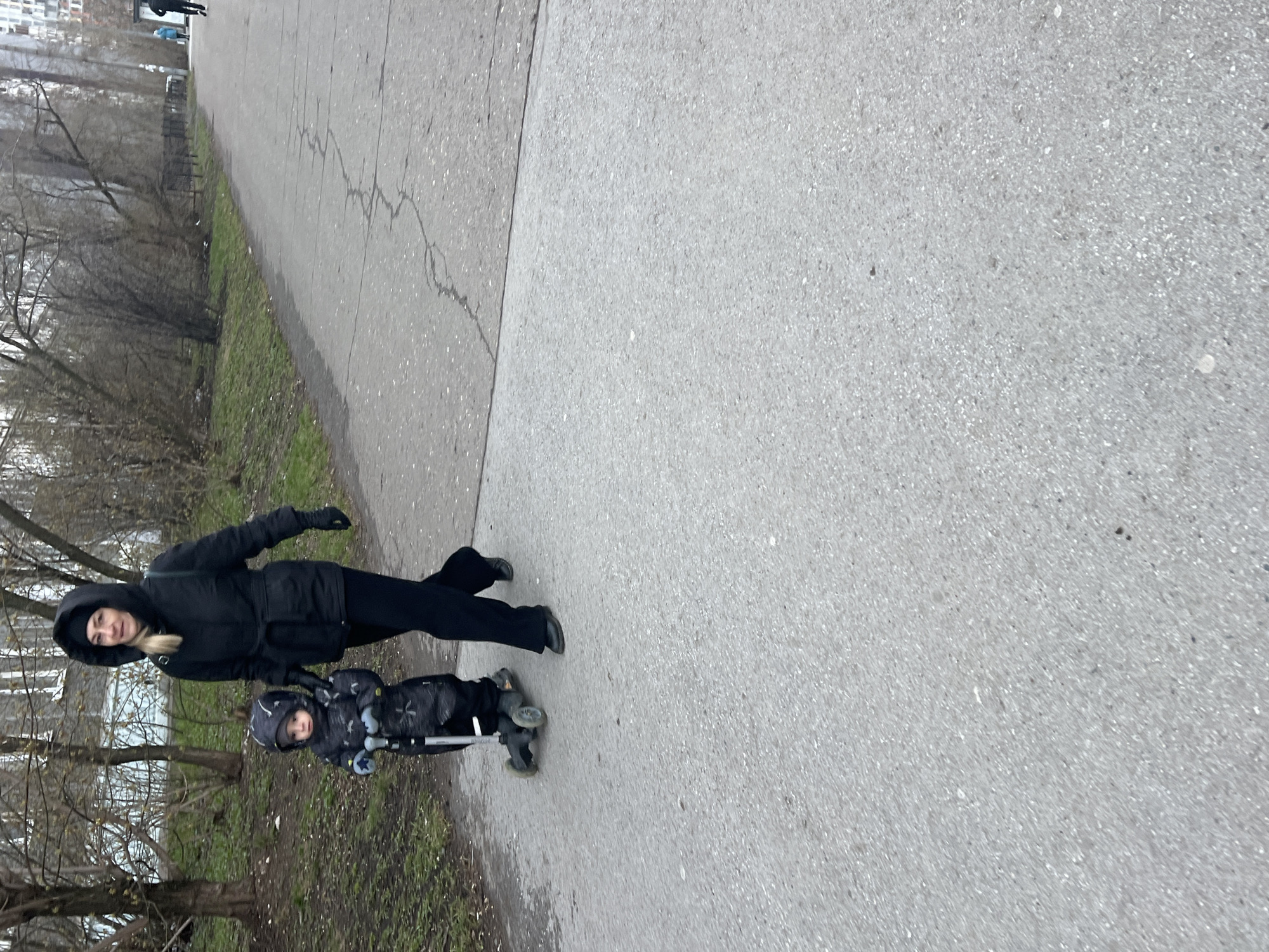 The image shows a woman and a child on a scooter on a sidewalk. The woman is wearing a black hooded jacket, black pants, and gloves. The child is wearing a dark patterned hooded jacket and pants, also on a scooter. They are on a paved sidewalk with grass and trees along the side. The scene appears to be set in a city park or a residential area. The location is Perm, Russia.