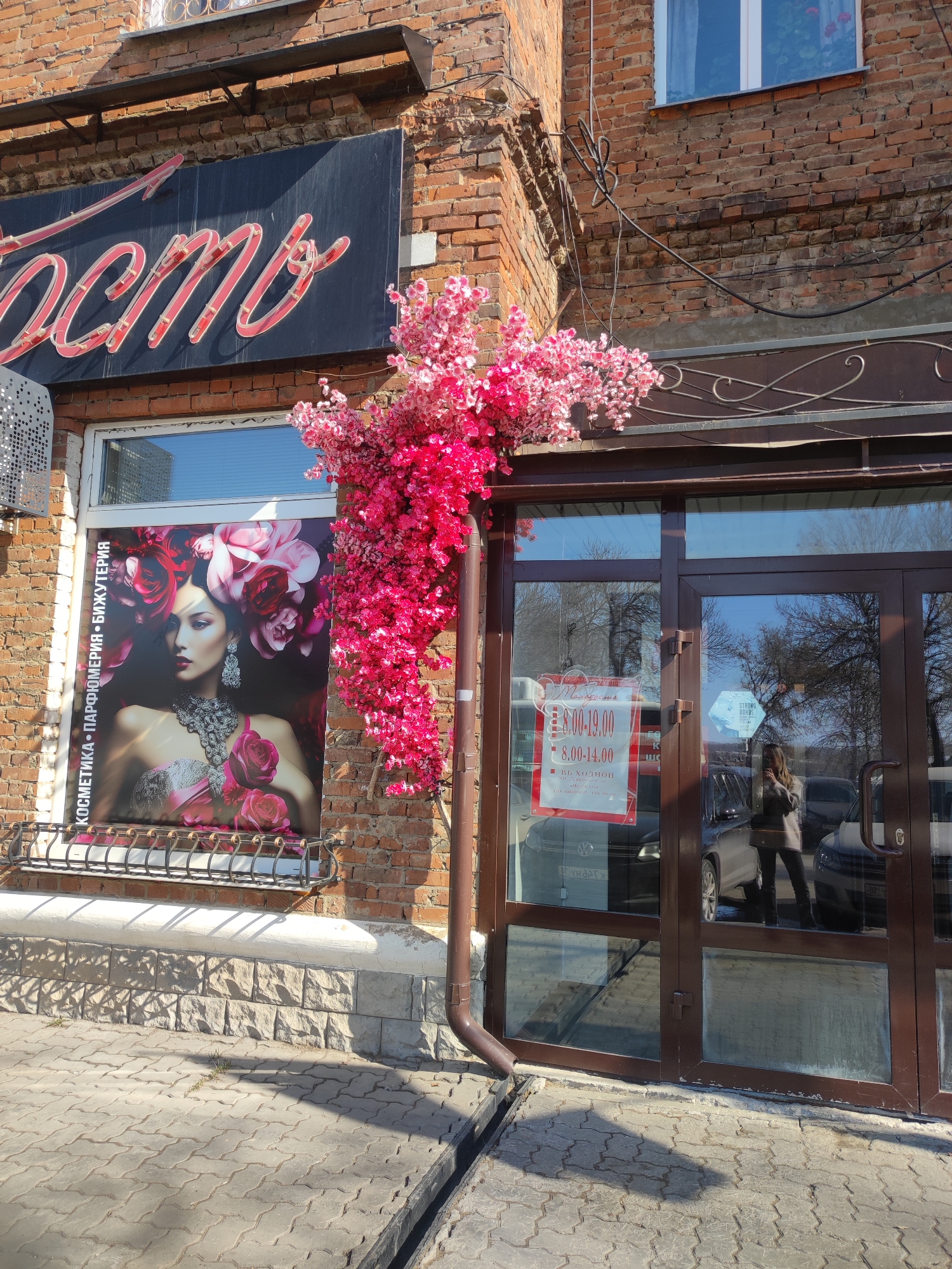 The photograph shows the exterior of a storefront in Votkinsk, Russia. The building is constructed of red brick with a stone base. Above the window, a black sign with script in a reddish-pink color is visible. The window to the left displays a large advertisement poster featuring a woman adorned with roses. To the right of the window, a cascade of bright pink artificial flowers is arranged. A brown metal rain gutter runs vertically down the corner of the building, partially obscured by the flowers. The entrance is a double glass door with a brown frame, reflecting the street scene, including cars and a woman standing outside wearing a light brown jacket and dark pants. A sign with the business hours is affixed to the door, showing the opening and closing times in red text. The sidewalk is paved with gray paving stones. The overall impression is that of a beauty salon or similar business.
