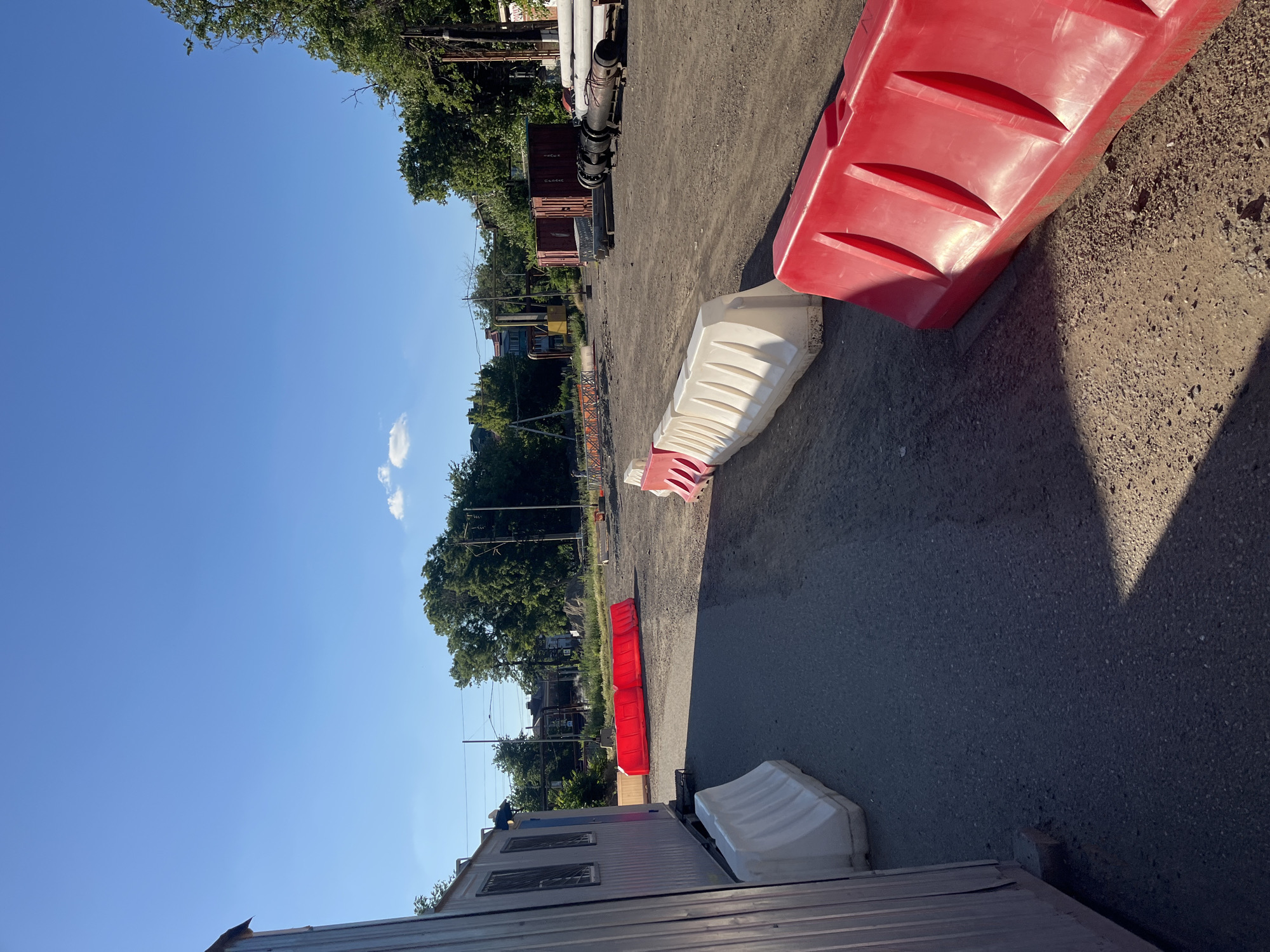 This image depicts a construction site under a clear blue sky. In the foreground, a grey corrugated metal structure is partially visible on the left. Several red and white plastic barriers are strategically placed across a gravel and asphalt surface, some creating pathways and delineating areas. In the midground, more red barriers are present. Further back, trees with lush green foliage line the horizon. Several shipping containers, one red and two grey, are situated in the middle distance. A stack of large cylindrical pipes, likely for construction, is visible on the right side of the image. In the background, buildings and electrical poles suggest an urban or suburban setting, possibly within the city of Krasnodar, Russia, though this cannot be definitively confirmed from the visual information alone. The scene appears to be an active construction or staging area.