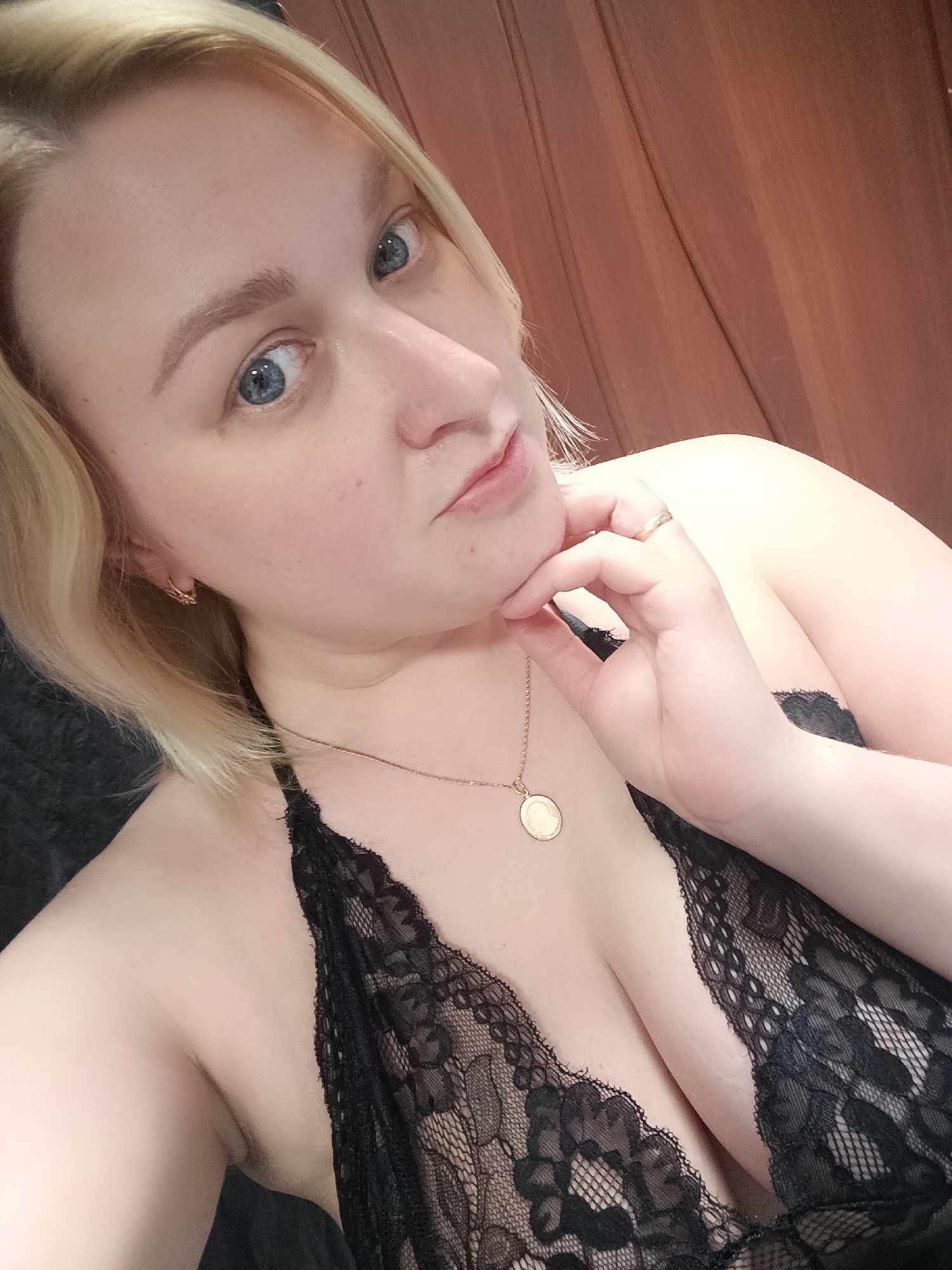A blonde woman with blue eyes is captured in a close-up selfie-style portrait. She has fair skin and short, light blonde hair. Her gaze is directed towards the camera with a slight pout on her lips, and her right hand is gently resting under her chin. She is wearing a black lace top or bralette, which reveals some cleavage. A delicate gold chain necklace with a circular, coin-like pendant is visible around her neck, and she has small gold hoop earrings. A plain gold band ring is on her left ring finger. The background consists of a warm, reddish-brown wooden wall, suggesting an indoor setting. The image is well-lit, highlighting her features. No visible text is present in the image. The implied location is Chaikovsky, Russia.