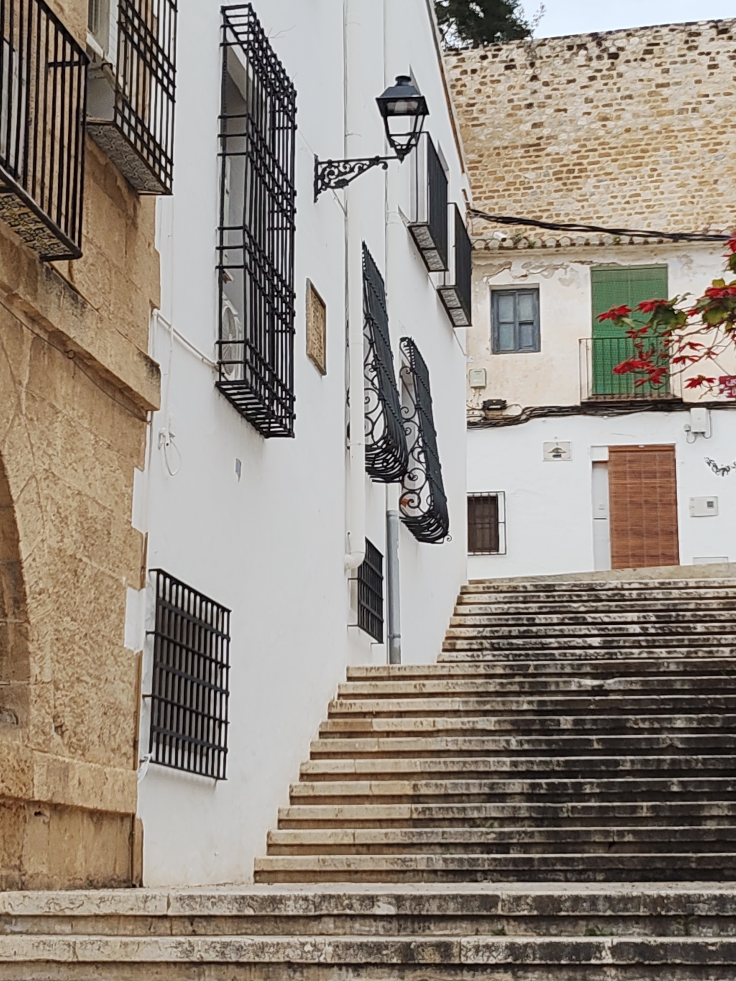 The image depicts a street scene in Dénia, Spain. The primary focus is a long set of stone stairs leading upwards alongside white-washed buildings. The architecture is characterized by black wrought-iron window grilles, and a vintage-style street lamp mounted on one building. Adjacent buildings have details like wooden doors and small balconies, as well as light brick wall. The image shows no people. The weather appears overcast.