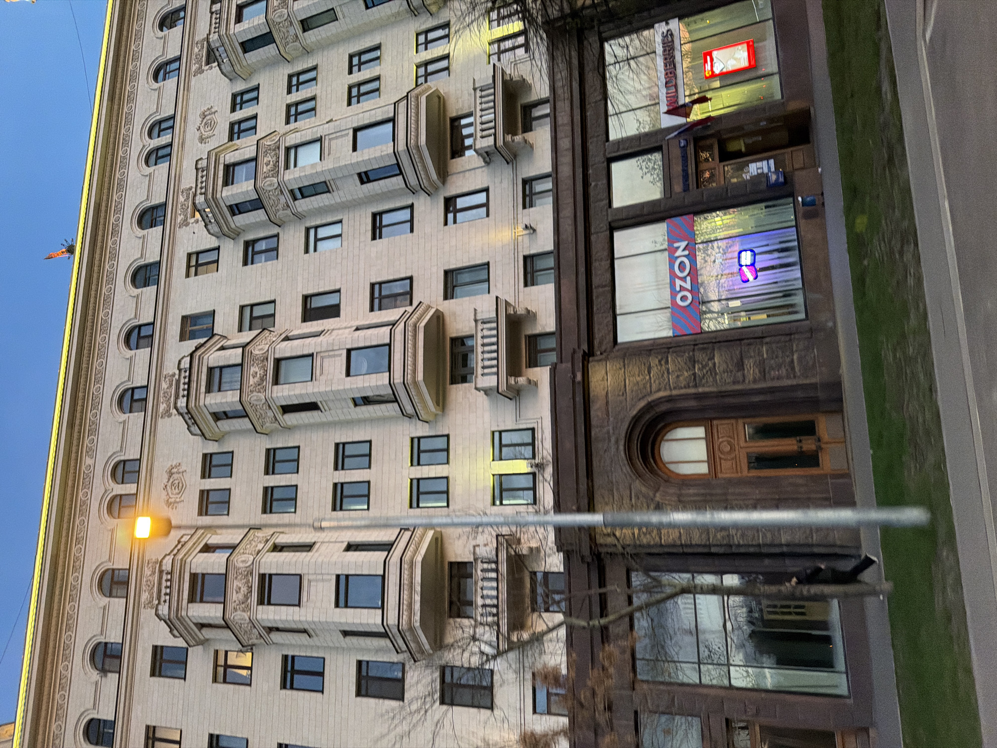 An eye-level, daytime, rotated medium shot shows a multi-story stone building in Moscow, Russia. The architecture style of the building is Stalinist, with numerous windows, balconies, and decorative elements on the façade. The building's exterior is predominantly light beige, contrasted with darker, possibly granite sections on the ground floor. Two large signs for "Wildberries" and "OZON" are visible, indicating commercial presence within the building. A blurred street lamp in the foreground casts an amber glow, and a portion of the street and a grassy area are visible. A person is seen lying on a tree branch in front of the building.