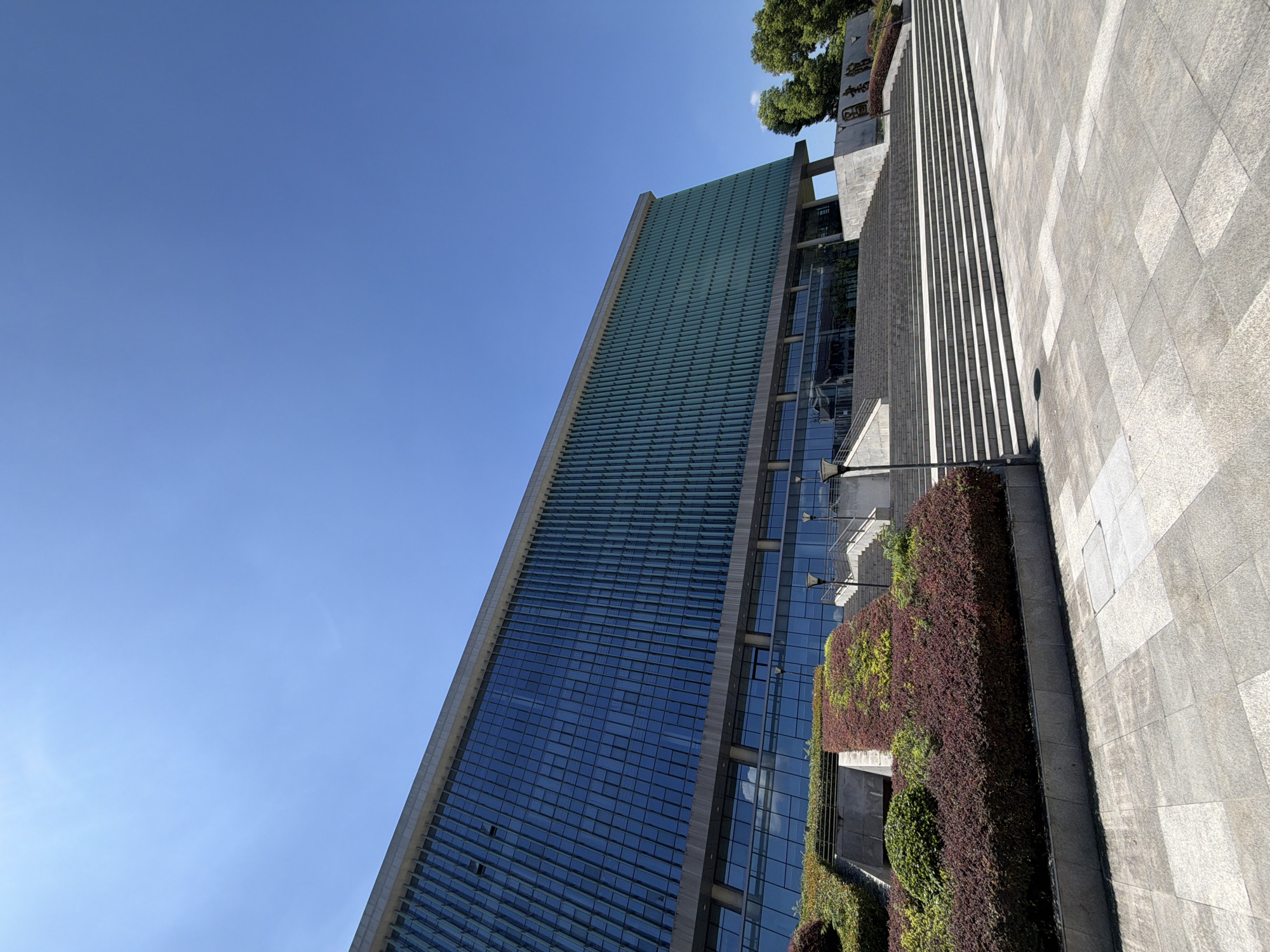 The image shows the exterior of a modern building with a glass facade and a wide set of stone stairs leading up to the entrance. The building has a long, horizontal design with multiple floors visible through the windows. The facade is composed of many vertical green fins, suggesting a modern architectural style. In front of the building, there are tiered landscaped areas with dark red and green foliage. The stairs are wide and made of light-colored stone. Several lampposts are visible along the stairs and on the plaza in front. The sky is clear and blue. To the right of the stairs, there appears to be a stone structure with Chinese characters inscribed on it, possibly a plaque or signage. The plaza in front of the stairs is paved with large, light-colored stone tiles. The scene is captured on a bright, sunny day. This building is the Jiangjin District Library in Jiangjin City, China.