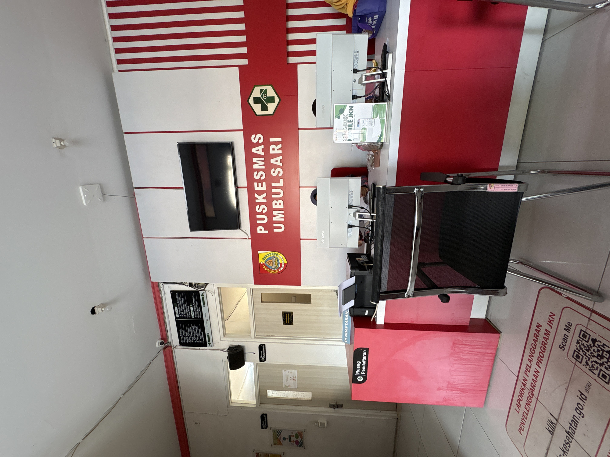 The image depicts the reception area of a public health center (Puskesmas) in Kebonsari, Indonesia. The walls are painted white with prominent red and white striped vertical accents. A large sign above the counter reads "PUSKESMAS UMBULSARI" in white lettering against a red background, accompanied by a green and white health service logo.
In the foreground, a red counter is visible, labeled "Ruang Pendaftaran" (Registration Room). A black office chair with chrome legs is positioned in front of the counter. On the counter, there are two computer monitors (one labeled Lenovo), a printer, and a green informational stand with text and a QR code. A purple bag with "HAPPY LOVE YOU" written on it sits to the right of the counter.
To the left of the counter, a door is labeled "Ruang Rekam Medis" (Medical Records Room). Further to the left, there is a sign with text and a QR code on the floor, reading "LAPORKAN PELANGGARAN PENYELENGGARAAN PROGRAM JKN" (Report Violations of the JKN Program Implementation).
The ceiling shows white with some exposed wiring and fixtures, including a light bulb and a security camera. The overall impression is a functional, albeit somewhat basic, administrative and service area of a local health facility.