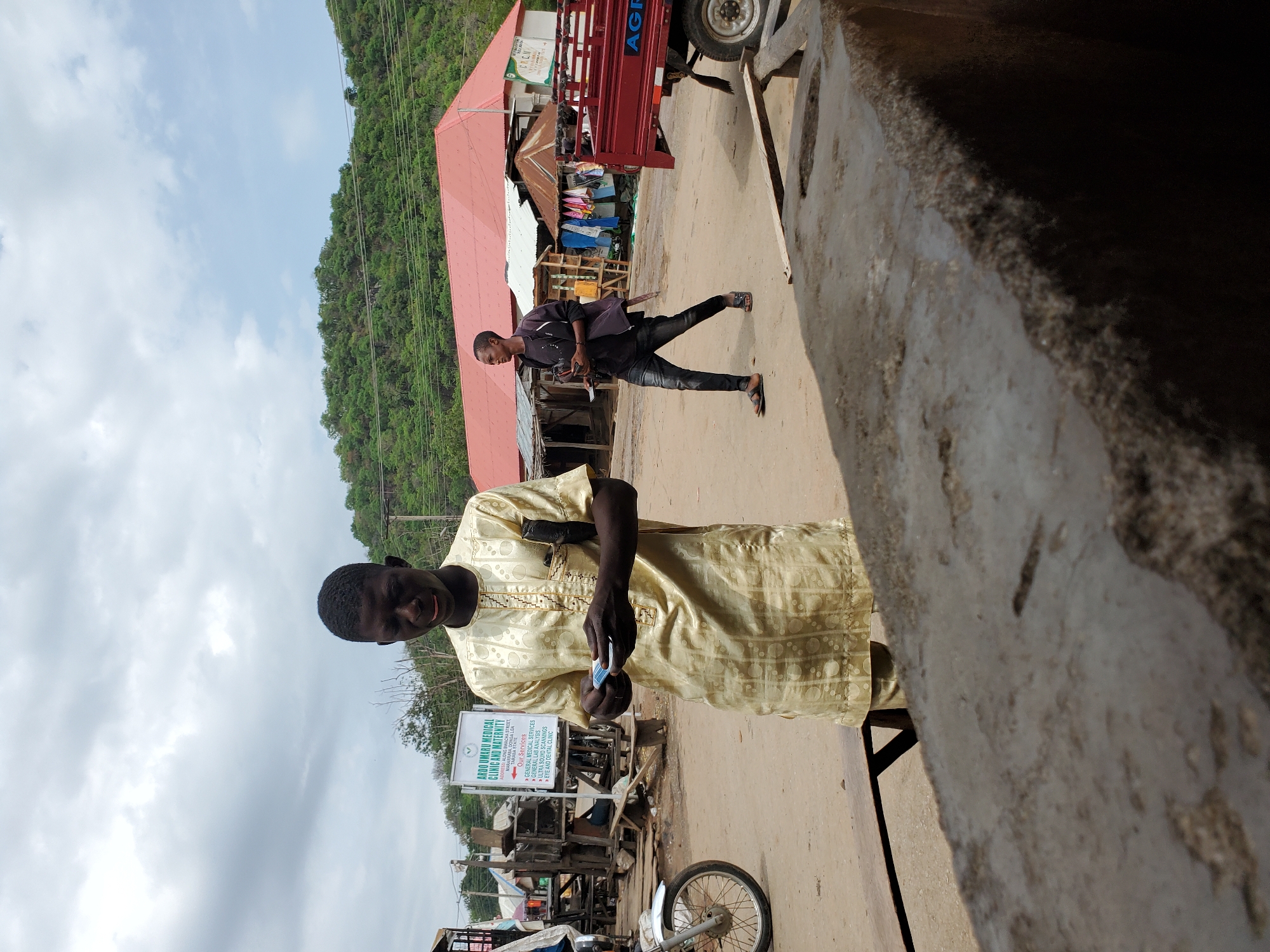 An outdoor scene in Mararaba, Nigeria, captures a bustling street with two men and several buildings. In the foreground, a man with dark skin, wearing a patterned light-gold traditional tunic, looks towards the viewer while holding a small rectangular object in his hands. He stands on an unpaved road, with a blurry, dark, textured wall occupying the right side of the frame.
Further back, another man in a dark jacket, dark shirt, and dark trousers walks away from the camera, glancing to his left. He wears sandals. The background features several informal market stalls or small shops with corrugated iron roofs. A red truck, partially visible on the right, has "AGF" emblazoned on its side.
To the left, a white sign with green lettering advertises "ARDO UMARU MEDICAL CLINIC AND MATERNITY," located "ALONG BIWACHA STREET, MARARABA." The sign lists services including "GENERAL MEDICAL SERVICES," "GENERAL LAB ANALYSIS," "ULTRA SOUND SCANNINGS," and "EYE AND DENTAL CLINIC." Behind the street and buildings, a prominent, lush green, tree-covered hill rises under a bright, partly cloudy sky. The overall scene depicts a typical daytime street environment, likely a market or commercial area, in Mararaba.