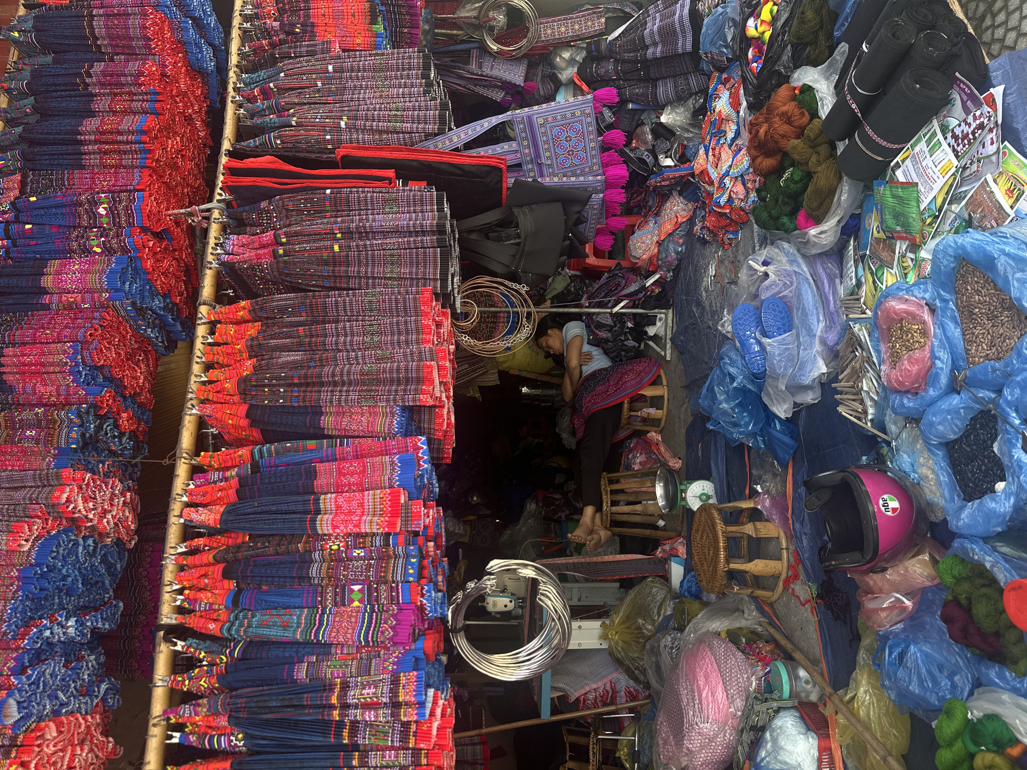 The image depicts a vendor at an apparent market in Púng Luông, Vietnam. Piles of vibrant, patterned textiles, predominantly red, blue, and black, are neatly stacked on the left and top left, occupying the majority of the frame. In the center, a woman is reclined on a small wooden stool, appearing to be sleeping, surrounded by an assortment of goods. These goods include bags, black fabrics, yarn, packaged goods, and seeds. A pink motorcycle helmet is situated amidst the collection. There are metal hoops/rings and a sewing machine on the left and front. The market is densely packed, with an abundance of merchandise on display.
