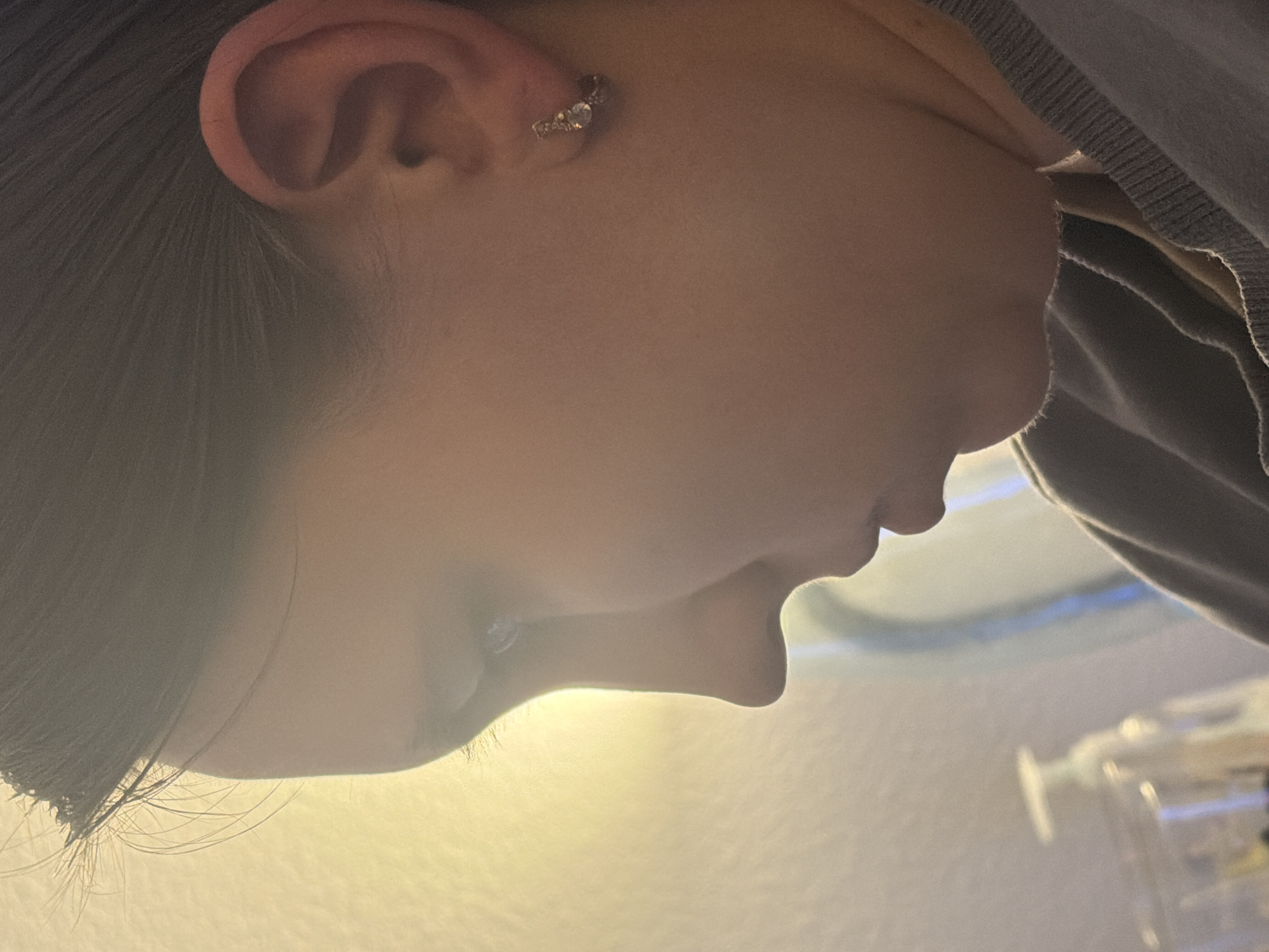 The image shows a close-up, profile view of a person's face, focusing on the ear and the side of their head. The person has dark hair and is wearing a small, stud-like earring with small stones. The lighting in the image is warm and appears to be coming from the left, casting a soft glow on the subject's skin and illuminating their features. The background is blurred and indistinct, with a light yellow hue. There are no visible elements in the image that suggest a specific location within the city of Portland, United States of America.