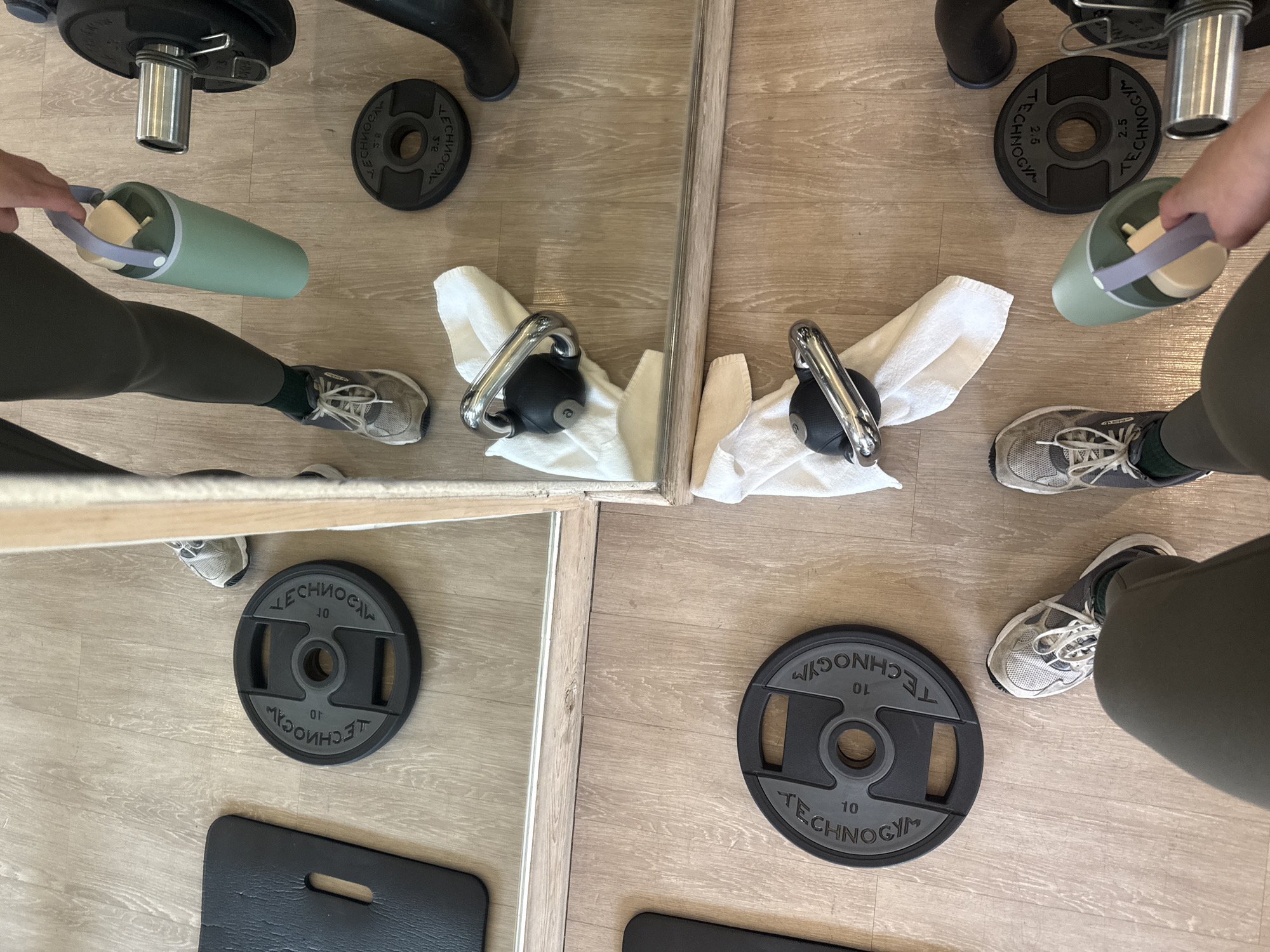 The image presents an overhead perspective of a gym setting, likely in Huixquilucan de Degollado, Mexico. The flooring appears to be a light-colored wood or laminate. In the center, a mirror divides the view, reflecting the items positioned in the gym.
There are two individuals, partially visible, each standing on either side of the mirror. They are wearing exercise attire, specifically olive green leggings and grey sneakers. One person is holding a light green water bottle with a grey handle.
On the floor are several pieces of gym equipment: two black kettlebells each resting on a white towel, and two black weight plates with the inscription "TECHNOGYM 10" and "TECHNOGYM 2.5" visible. Additionally, a black exercise mat is located in the lower left corner.
The equipment and individuals are situated in proximity to the mirror, creating a mirrored reflection of the scene.
