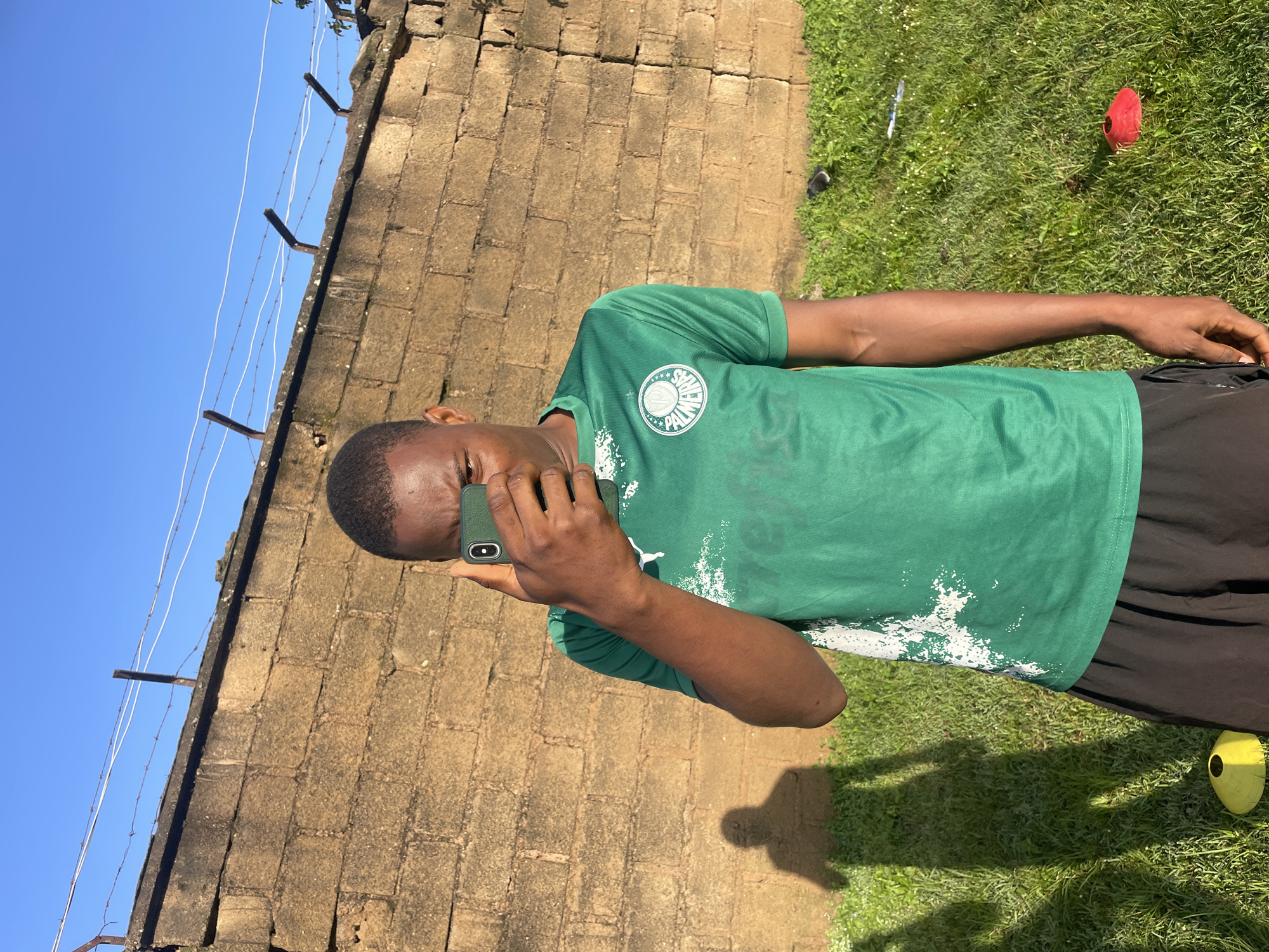 A young Black male, wearing a green soccer jersey with a white emblem and black shorts, stands outdoors in front of a textured brick wall. He is holding a green smartphone up to his face, as if taking a selfie or recording a video. The bright blue sky is visible above the wall, which is topped with barbed wire. The ground is covered in green grass, with yellow and red cones scattered on it, suggesting a sports or training area. The sun casts a long shadow of the man onto the grass. The scene appears to be in Jos, Nigeria, given the context provided.