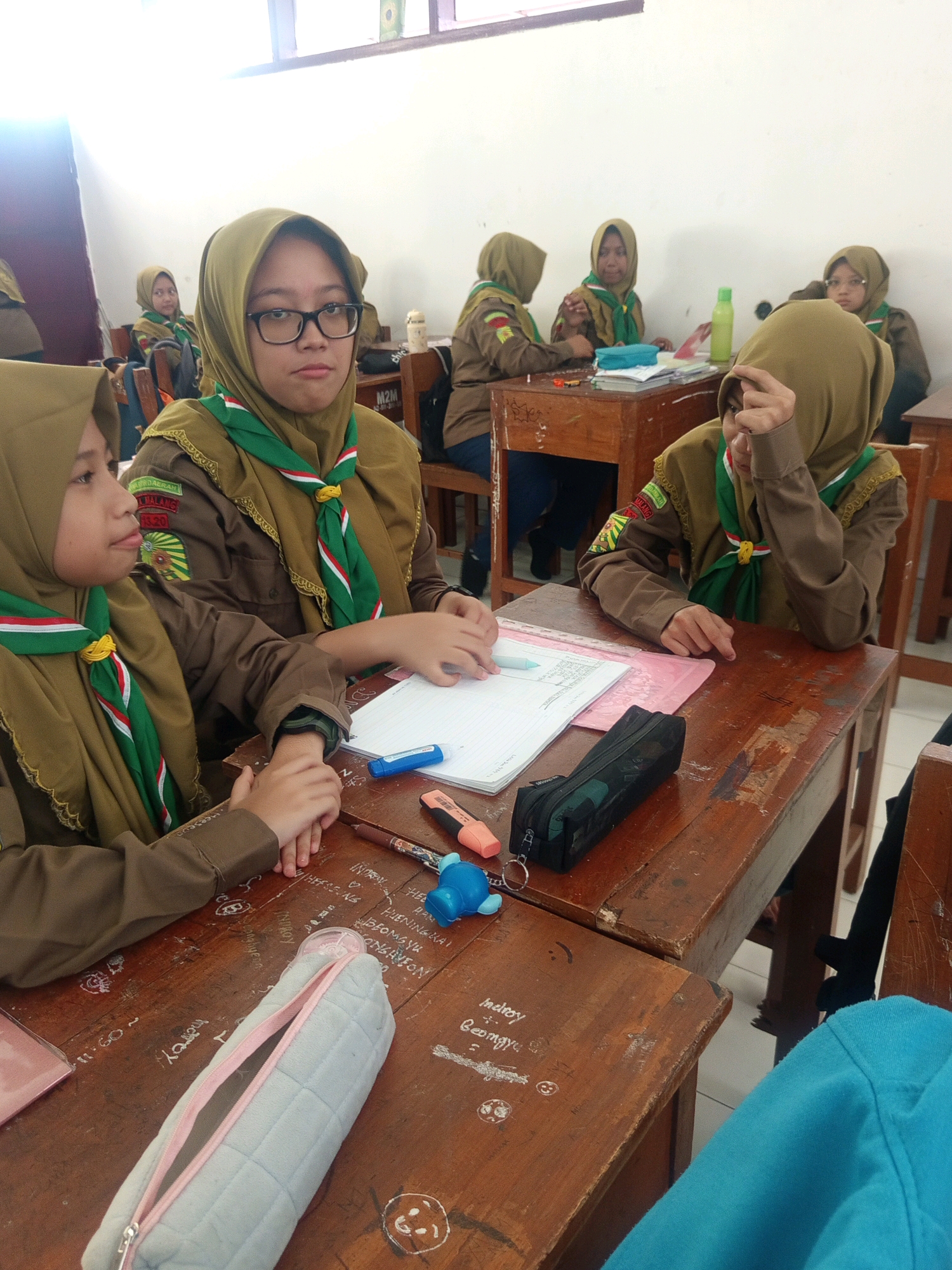 The image shows a classroom setting with several young female students wearing scout uniforms. They are seated at wooden desks, some of which are decorated with graffiti. The students appear to be engaged in schoolwork, with notebooks, pens, and other stationery visible on the desks. One student in the foreground is looking directly at the camera, while others are focused on their tasks or interacting with each other. The classroom appears to be in Malang, Indonesia, as indicated by the badge on one of the students' uniforms which reads "KWARTIR DAERAH JAWA TIMUR MALANG 33.20". The lighting suggests it is daytime, possibly an indoor activity or event.