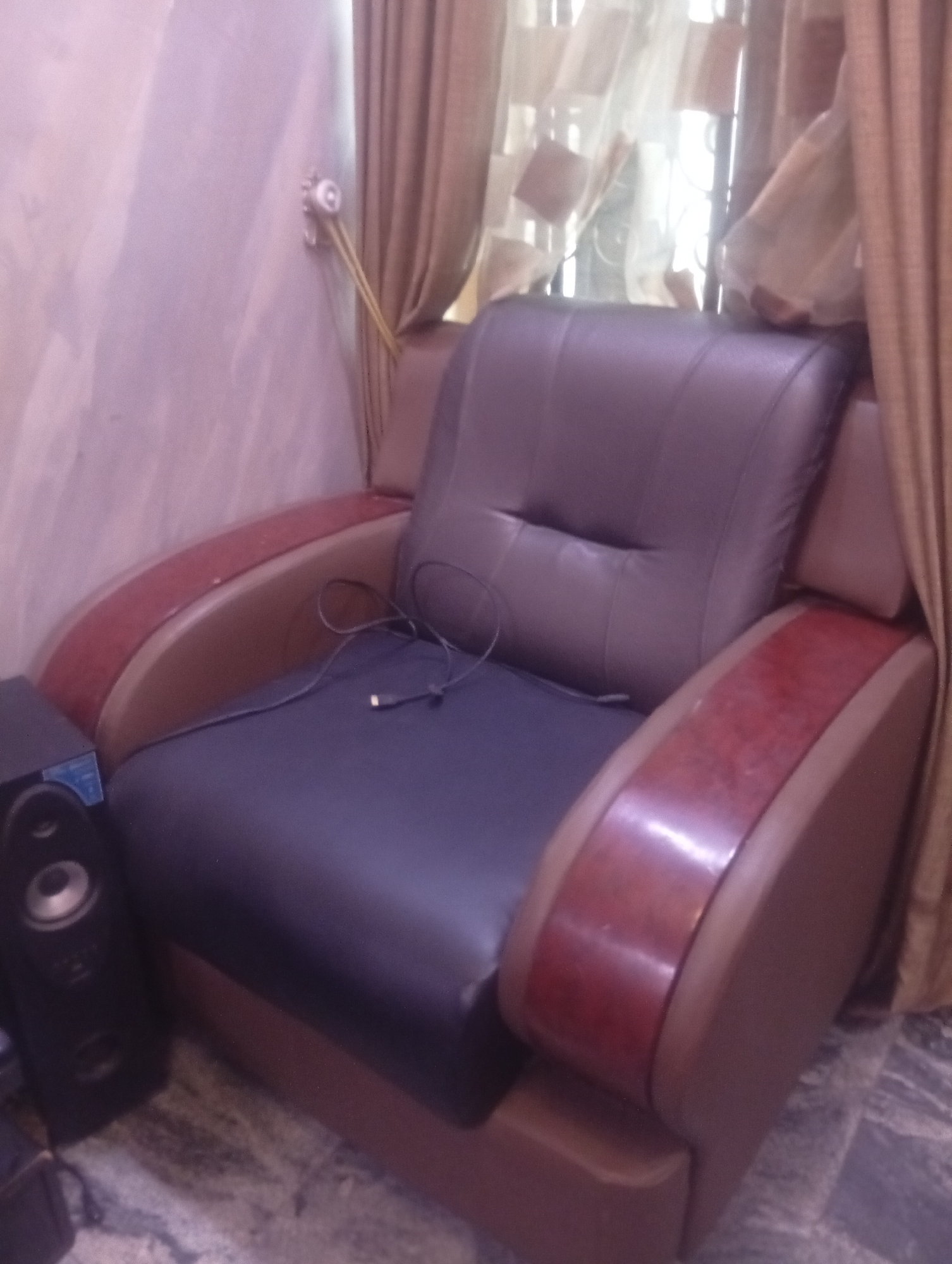 The image shows a dark blue leather armchair with brown wood armrests in what appears to be a domestic setting. A black speaker is positioned to the left of the chair. A black USB cable is on the chair. Curtains are draped behind the chair. The image was taken in Aviation Quarters, Nigeria.