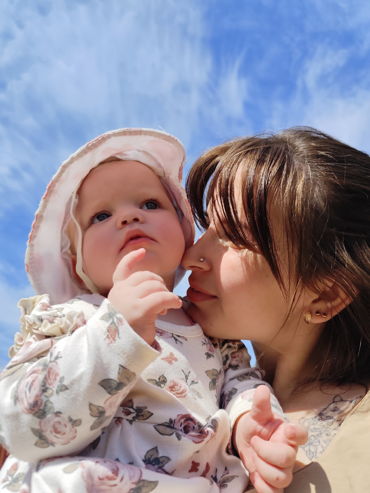 A woman holds a baby in her arms against a backdrop of a blue sky with wispy clouds. The baby is wearing a bonnet and a floral patterned outfit. The woman is close to the baby, and has dark hair and fair skin. The location in the city of Фоки, Russia cannot be determined from the image.