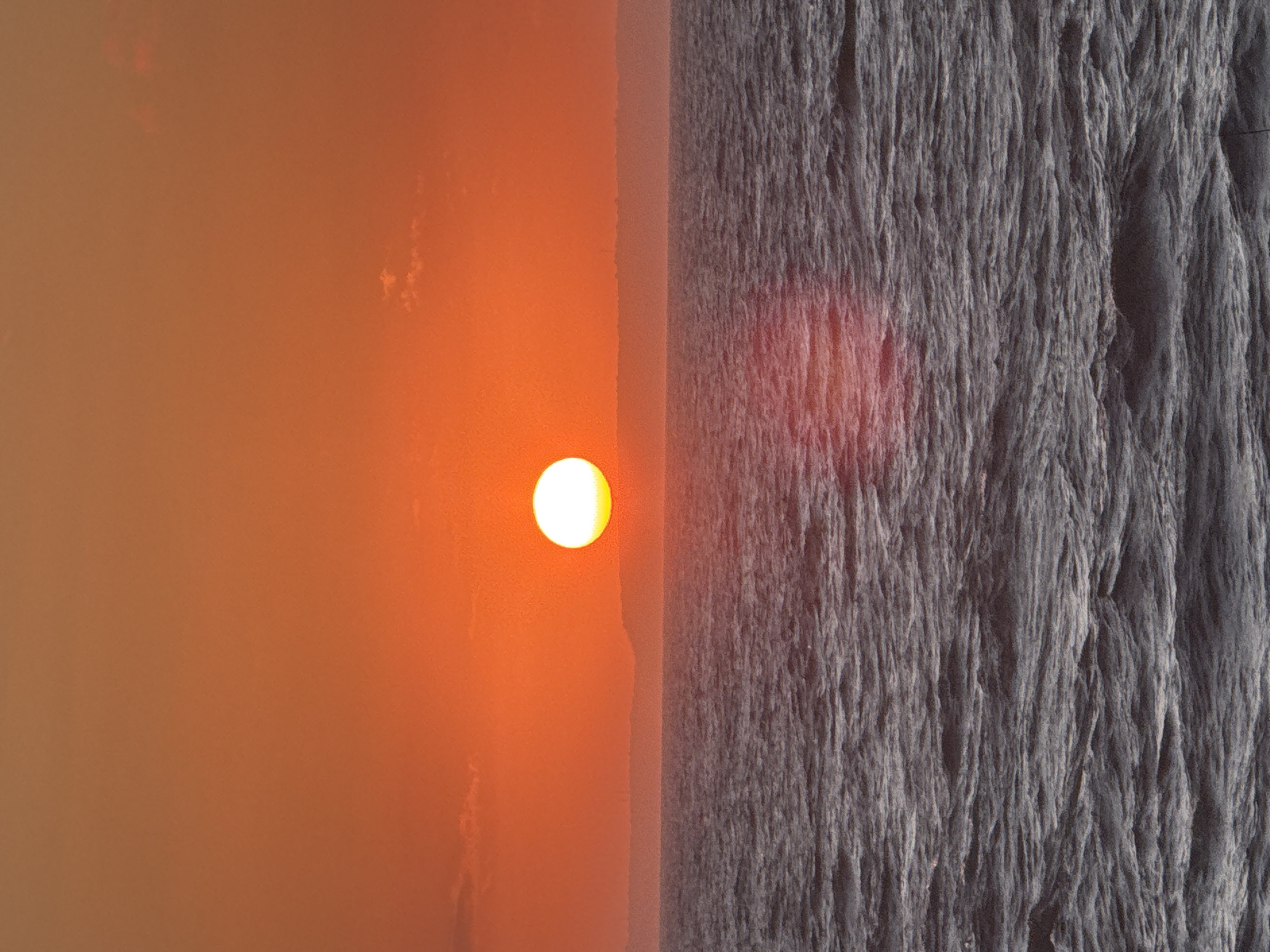 This media file captures a vibrant sunset or sunrise over a body of water in Kuta Selatan, Indonesia. The image is divided horizontally by a clear horizon line. The left side is dominated by a warm, orange-red sky, heavily diffused and hazy, suggesting atmospheric particles like dust or smoke, or perhaps thick humidity. A bright, circular sun, appearing almost white at its core due to its intensity, is positioned centrally in the left half, just above the horizon.
The right side of the image depicts a textured surface, likely the ocean, with strong patterns resembling rough waves or currents, possibly viewed from an elevated perspective or through a unique filter. This water surface is predominantly dark grey and black, with noticeable vertical striations and ridges that create a sense of movement and depth. A faint, reddish glow, mirroring the color of the sky and the sun, is visible on the water surface directly beneath the sun, indicating a reflection. There are no visible people, artificial objects, or text. The overall atmosphere conveyed is one of natural beauty and dramatic light.