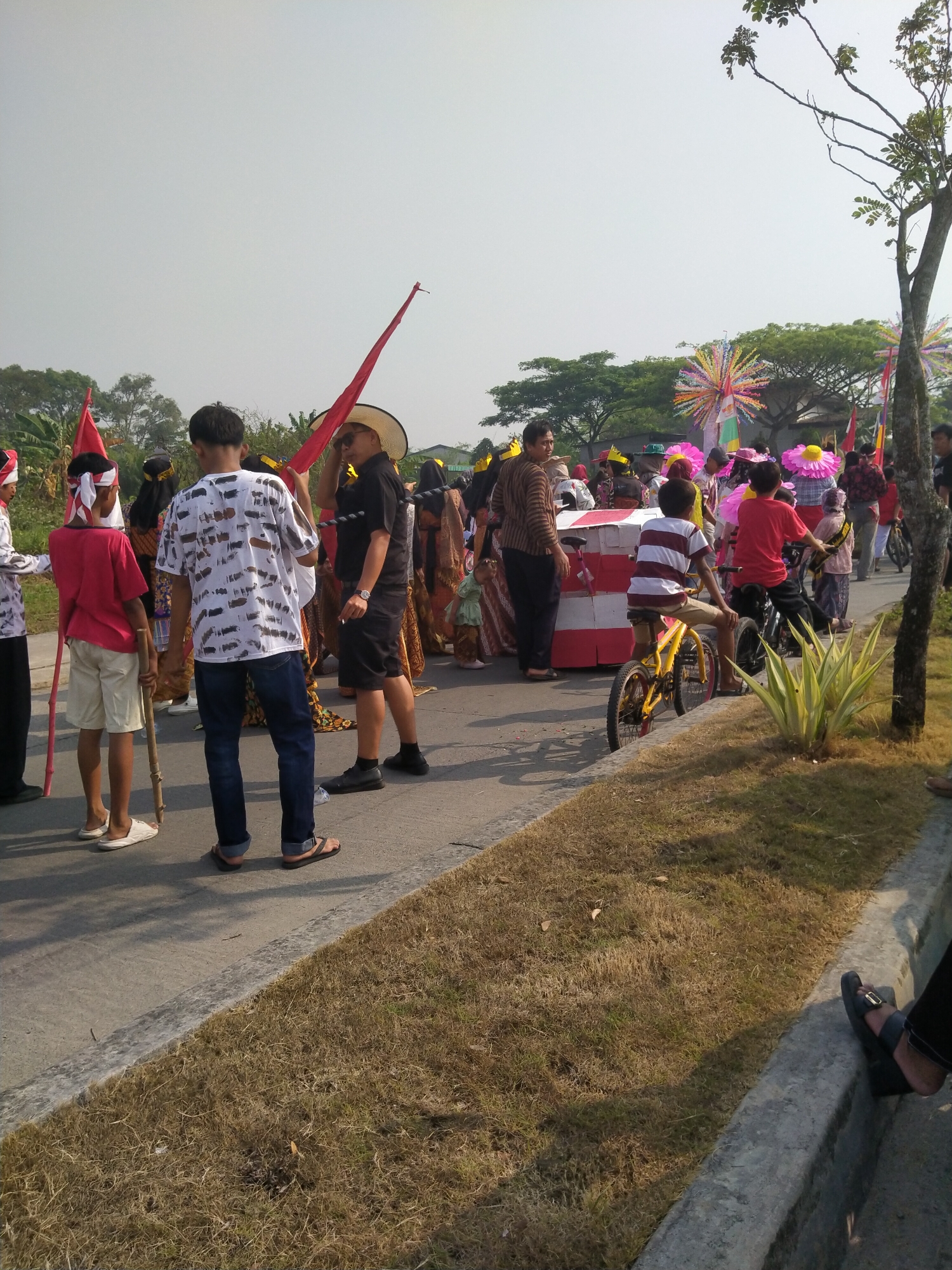 This image captures a street scene in Cisoka, Indonesia, likely during a local event or parade. In the foreground, two boys are on bicycles. The boy on the left is wearing a striped shirt and shorts, while the boy on the right is in a red t-shirt and dark pants. They are on a paved road, with dry grass and a concrete curb to their right.
Behind them, a diverse group of people are gathered, some in traditional attire. A man in a black shirt and straw hat stands holding a red flag. To his left, a young boy in a red shirt and white headband holds a wooden stick. Further back, more individuals are visible, some carrying flags or decorated objects, suggesting a celebratory atmosphere. The background features trees and a bright, hazy sky. The overall scene conveys a sense of community gathering and cultural participation.