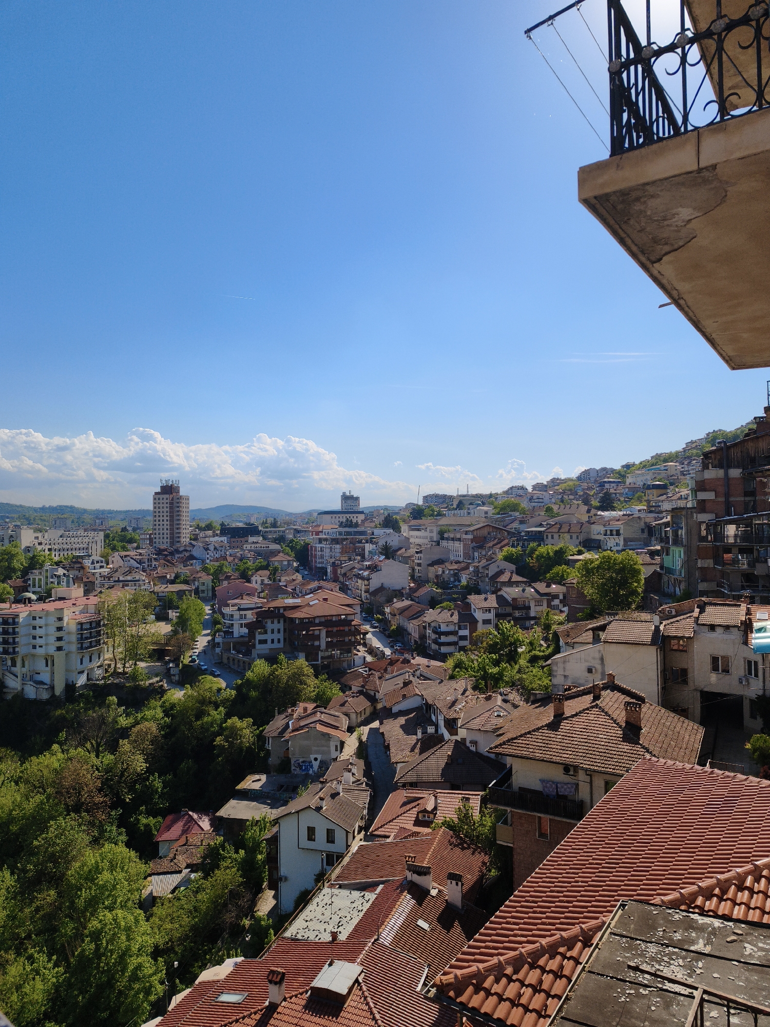 The image shows a daytime aerial view of Veliko Tarnovo, Bulgaria. The sky is blue with scattered clouds. The cityscape is dominated by buildings with red-tiled roofs and various structures, including a high-rise building in the distance. Green trees are interspersed throughout the residential areas. A balcony with a wrought-iron railing is visible in the upper right corner of the frame, suggesting the photograph was taken from a high vantage point, likely a balcony or rooftop.