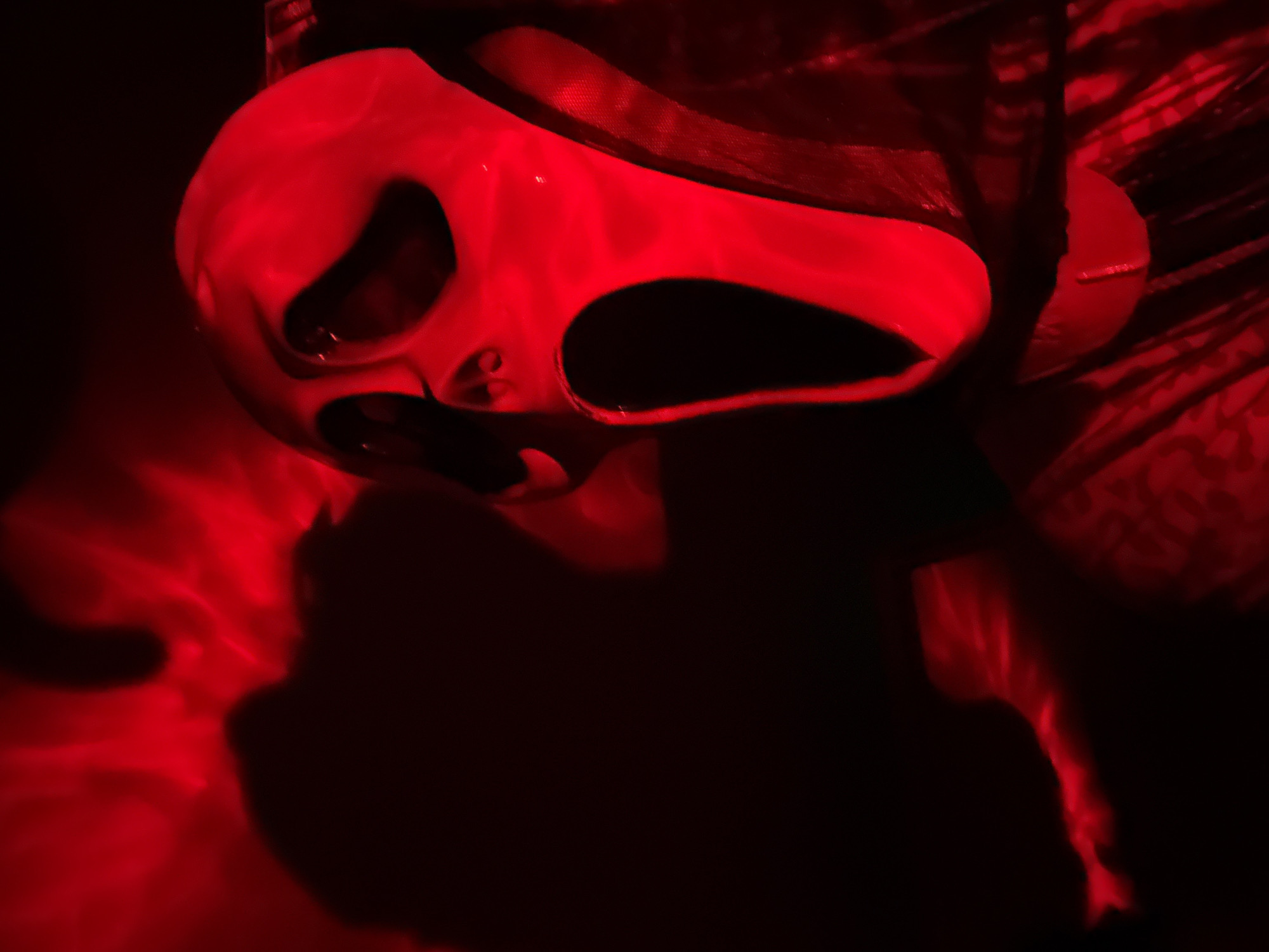 A white, mask-like object with hollow eyes and mouth is illuminated by red light. The mask is positioned in the upper center of the frame, casting shadows in the lower portion of the image. The background is dark and indistinct, suggesting an indoor setting with low ambient light. A textured, possibly cloth-like material is visible behind and to the right of the mask. Location information is unavailable.