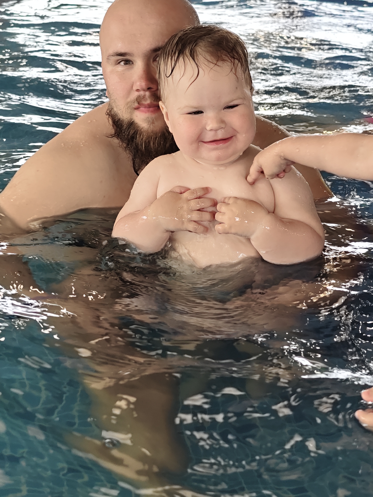 A bald man with a beard is waist-deep in a pool, holding a baby in front of him. The baby is smiling and has their hands clasped together. A child's hand is reaching towards the baby. The pool has a blue tiled bottom. The location may be a swimming pool in the city of Votkinsk, Russia, but this cannot be confirmed solely from the image.