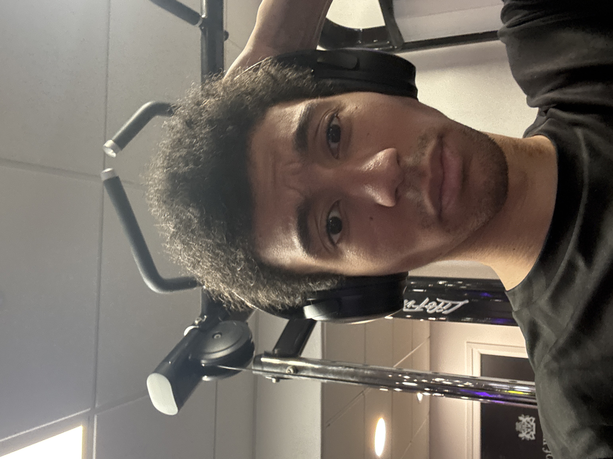An indoor, close-up selfie-style portrait captures a man in what appears to be a gym setting in London, United Kingdom. The subject, a man with dark, curly hair and light facial stubble, looks directly at the camera with a pensive or slightly furrowed expression. He is wearing black over-ear headphones and a dark (likely black) t-shirt.
Behind him, various pieces of gym equipment are visible, featuring metallic elements and dark padding. To the left, part of a gym handle or bar is seen, and a larger black panel stretches across the background. On this panel, the brand name "Life Fit" is distinctly visible in a white, stylized font. Below "Life Fit," a partially obscured crest or emblem-like logo can also be seen. The lighting suggests an indoor environment, likely from overhead artificial sources, illuminating the scene brightly. The overall impression is that of a workout session or a pause during one.