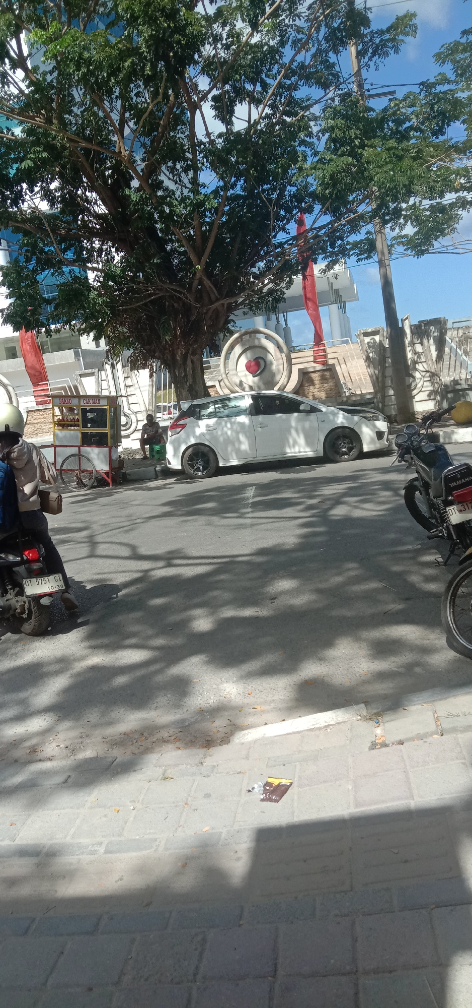 An outdoor, eye-level shot captures a street scene in Kendari, Indonesia, under a clear blue sky. A large tree with lush green foliage dominates the left side of the frame, its branches casting dappled shadows on the asphalt road. In the middle ground, a white hatchback car is parked, its sleek design contrasting with the rustic charm of a nearby food cart labeled "BAKSO CAK MAN." A person is seated on a green plastic chair beside the cart, seemingly engaged in conversation or waiting. A second individual, wearing a helmet, is seated on a motorcycle, while another motorcycle is parked to the right of the car, displaying a license plate that reads "DT 3172." The background reveals a modern building with a glass facade, interspersed with red banners and an artistic circular monument featuring a red heart. The street is paved with interlocking blocks, and scattered leaves suggest a recent breeze. The overall impression is one of everyday life unfolding in a vibrant urban setting.
